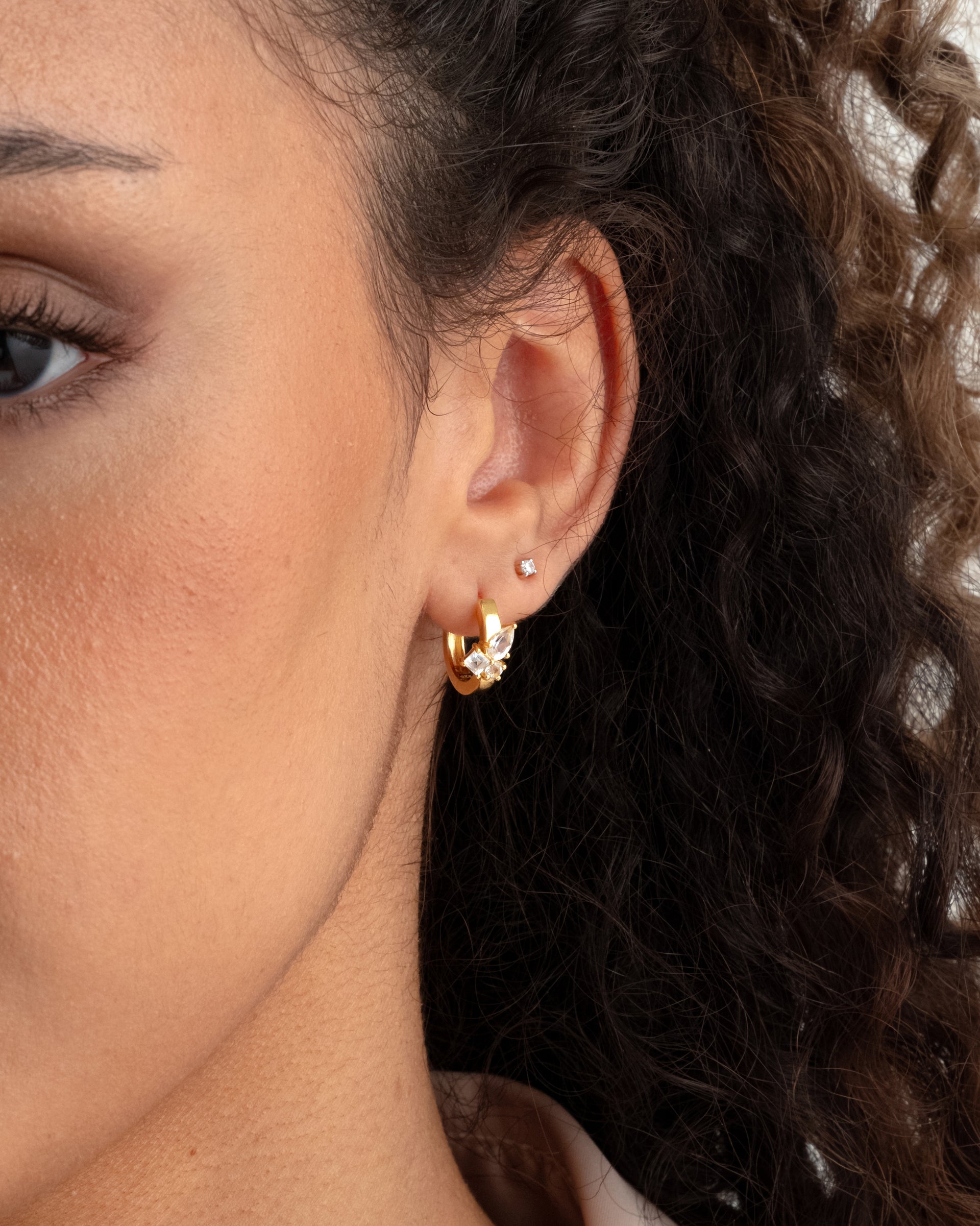 A close-up shot of a model wearing the gold Pam Hoops, adorned with white topazes, and a small crystal stud earring.