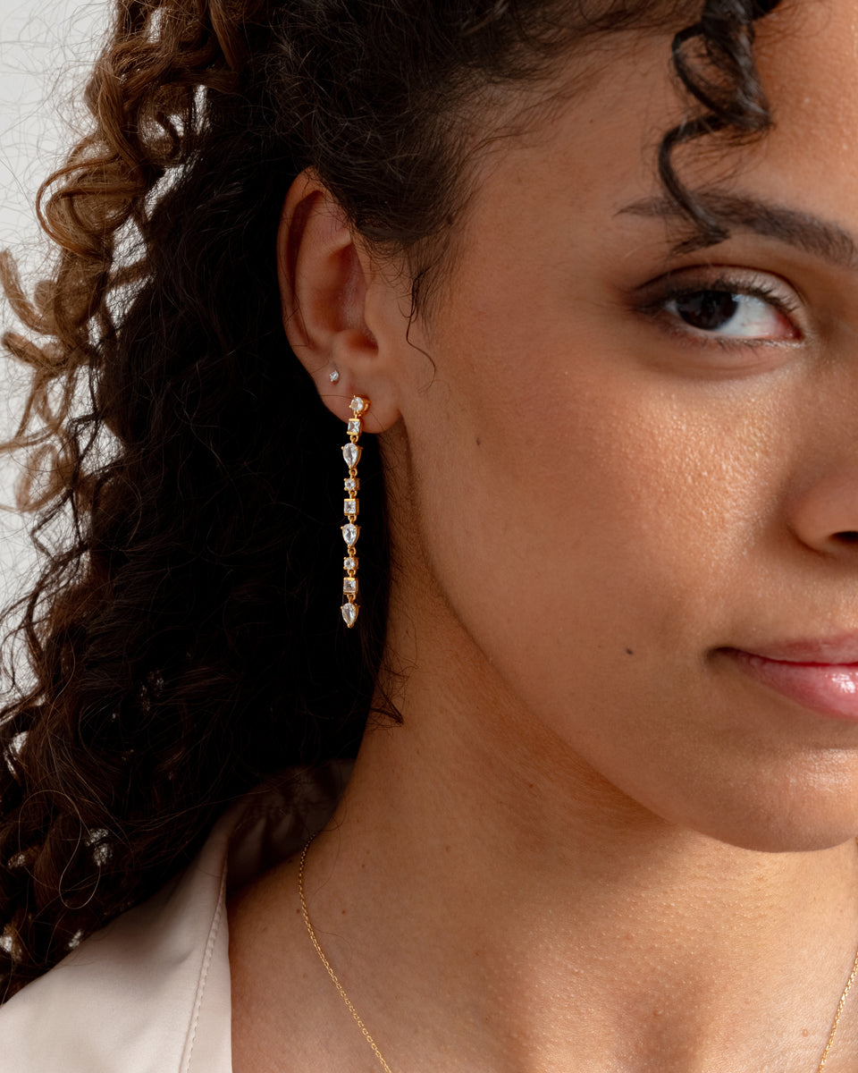 A close-up shot of a model wearing the Pam Long Earrings in gold, a dangling design featuring a cascade of sparkling white topazes in various shapes.