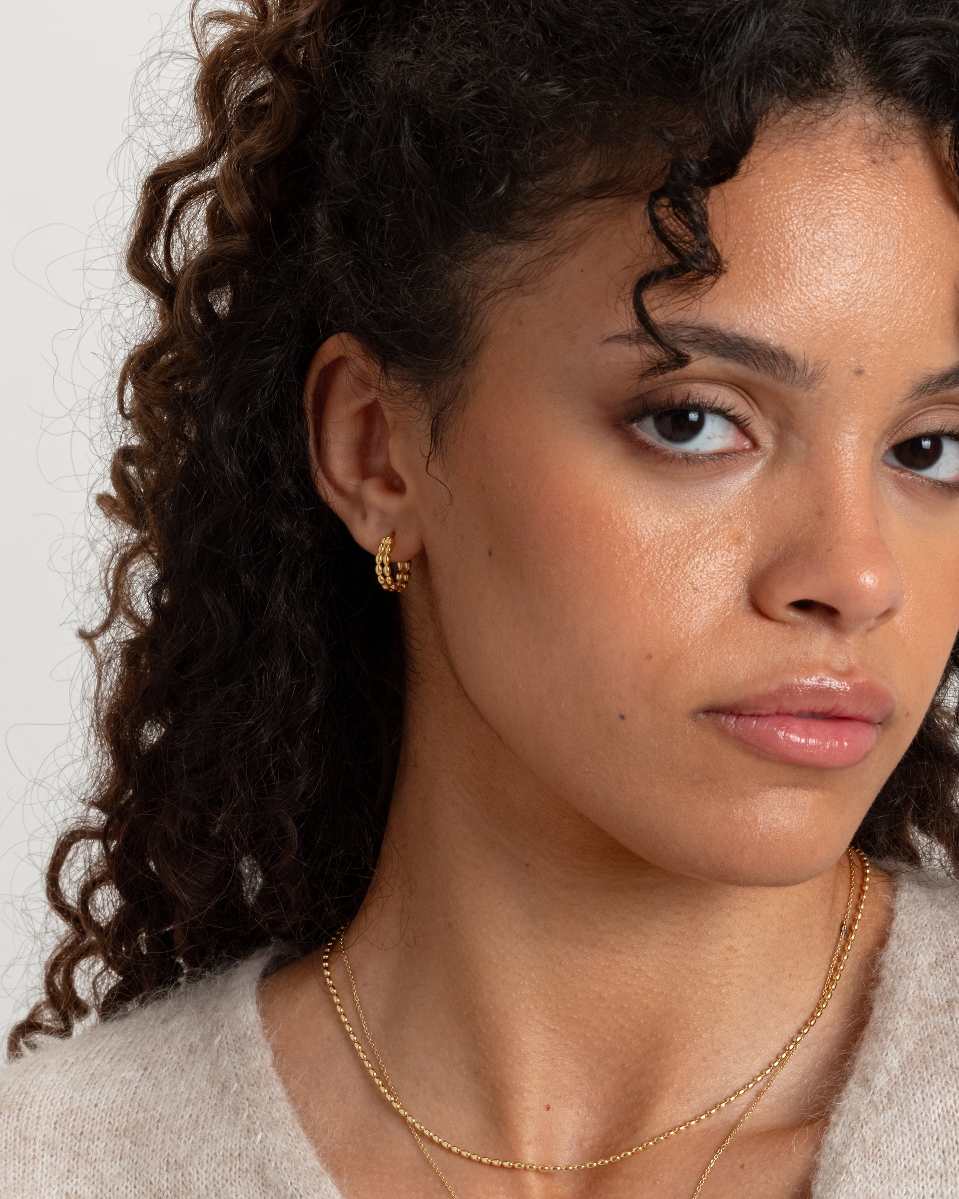 A close-up of a model wearing the Lunetta Earrings in gold, featuring their unique design of interconnected ovals, paired with two layered gold chain necklaces.