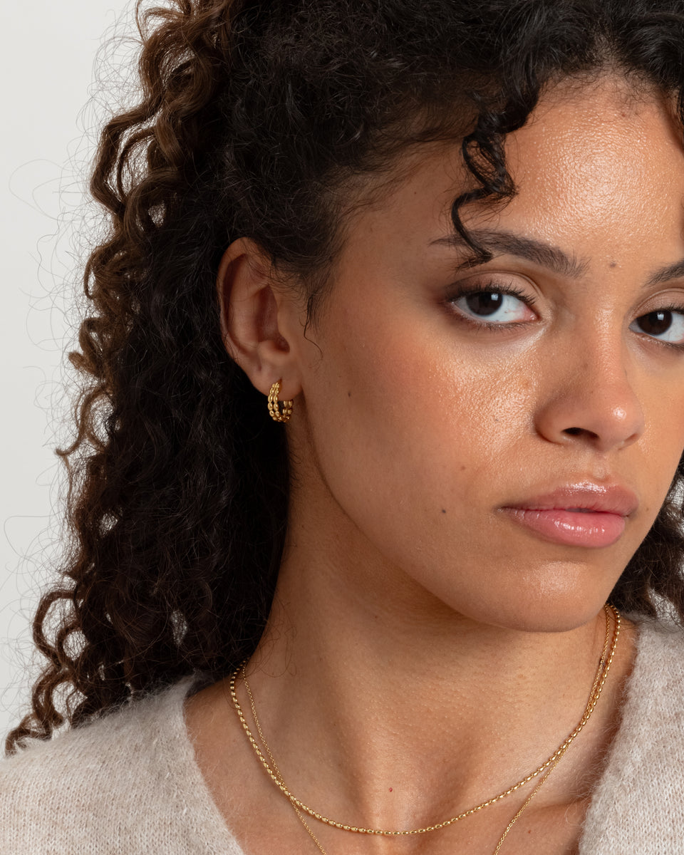 A close-up of a model wearing the Lunetta Earrings in gold, featuring their unique design of interconnected ovals, paired with two layered gold chain necklaces.