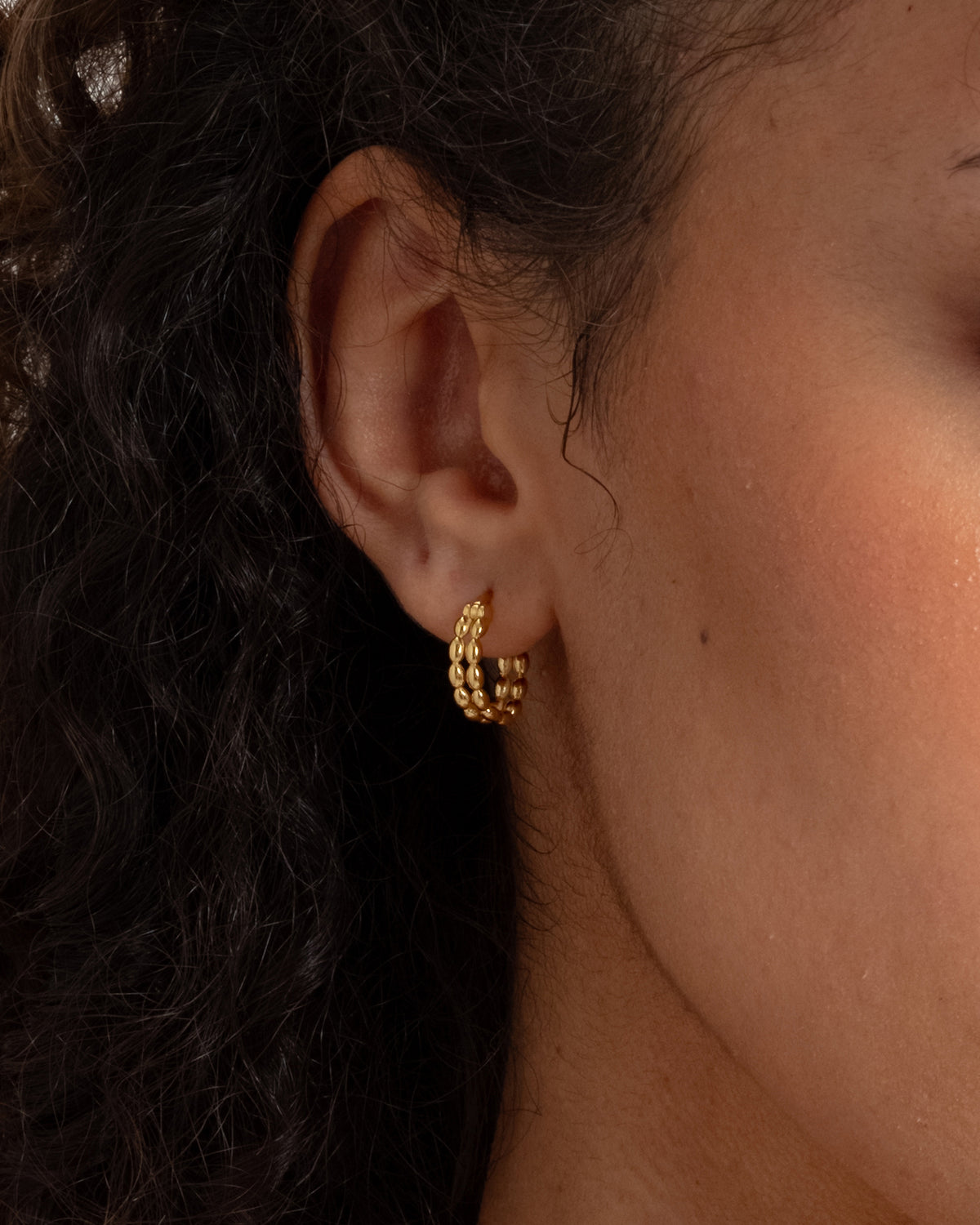 A close-up shot of a model wearing the Lunetta Earring in gold, highlighting its unique design of two parallel circles linked together by a pattern of small ovals.