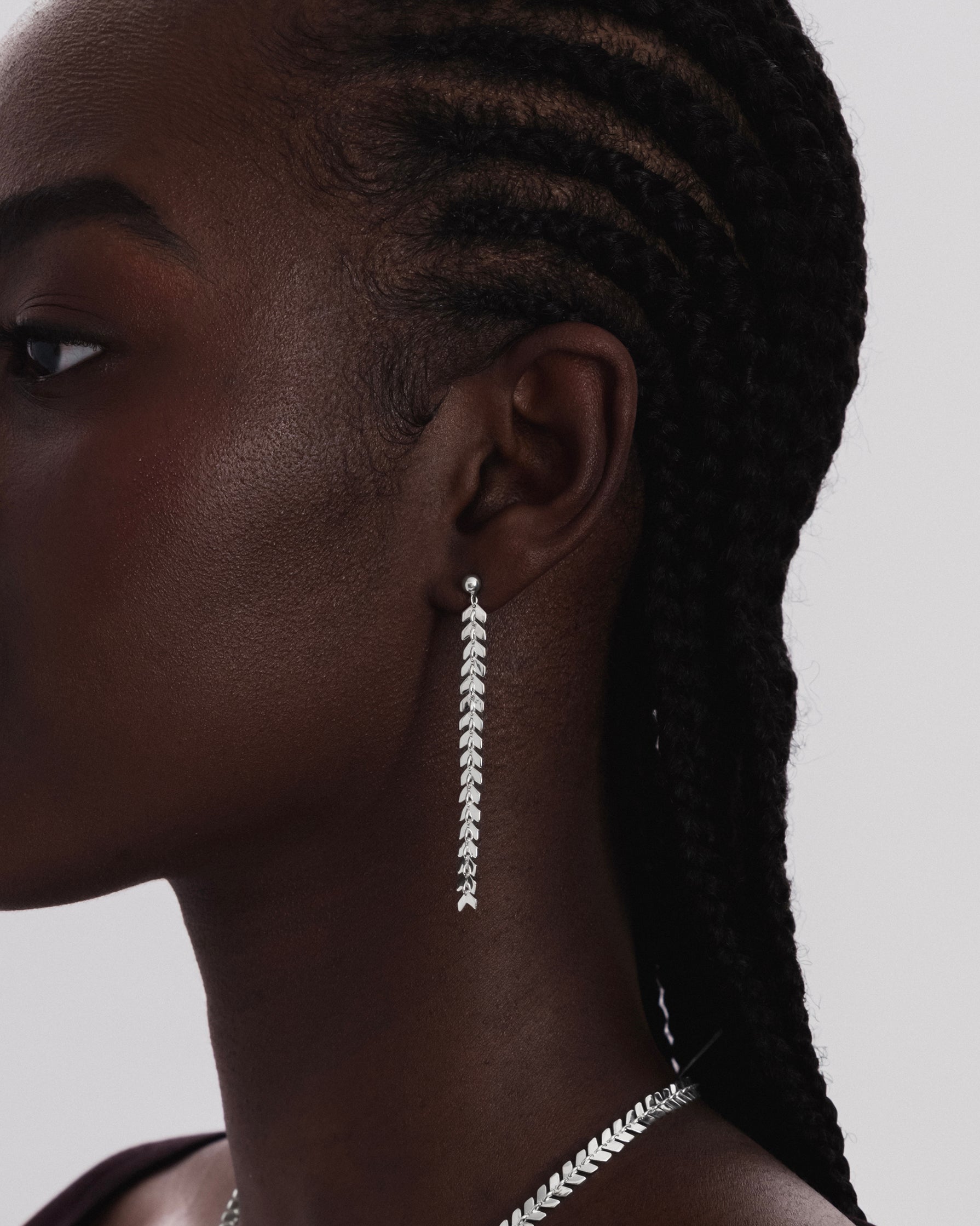 A close-up shot of a model wearing the Layers Olivia Earrings in silver, which feature a long, dangling chain of small leaf-like shapes. A matching necklace is also visible.