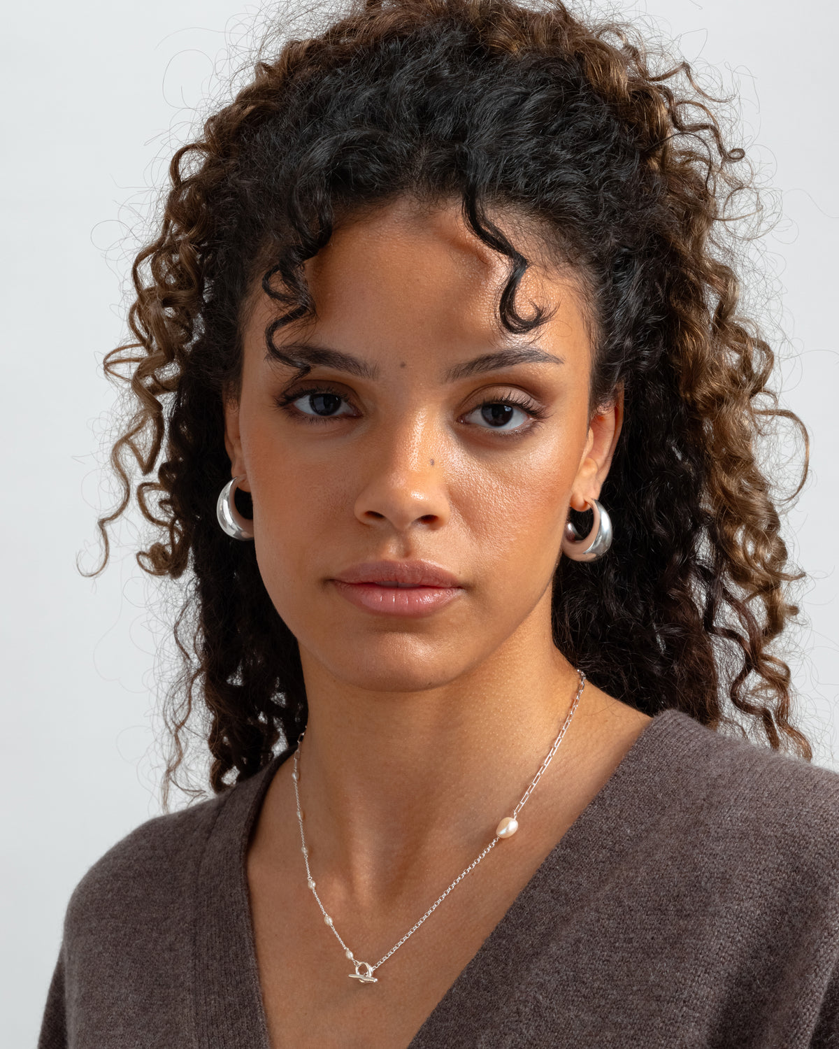 A close-up of a model wearing the Bolded Earrings in silver, a pair of chunky hoops, and a delicate silver necklace with pearls.