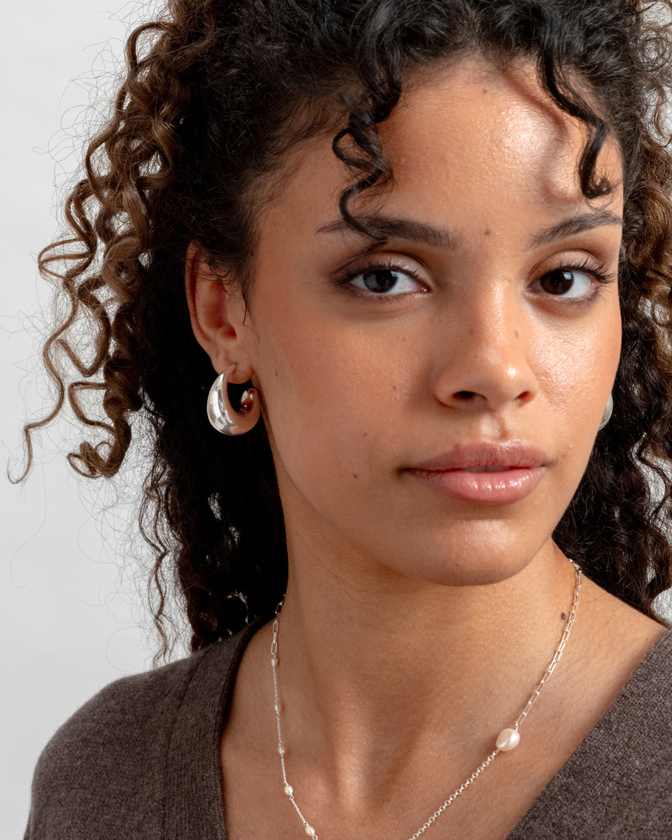 A close-up of a model wearing the Bolded Earrings in silver, featuring a chunky, unique hoop shape, along with a fine silver chain necklace with small pearl details.