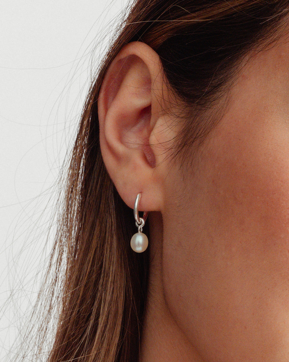 A close-up of a model wearing the Treasure Pearl Hoop in silver, featuring a delicate hoop with a single dangling freshwater pearl.