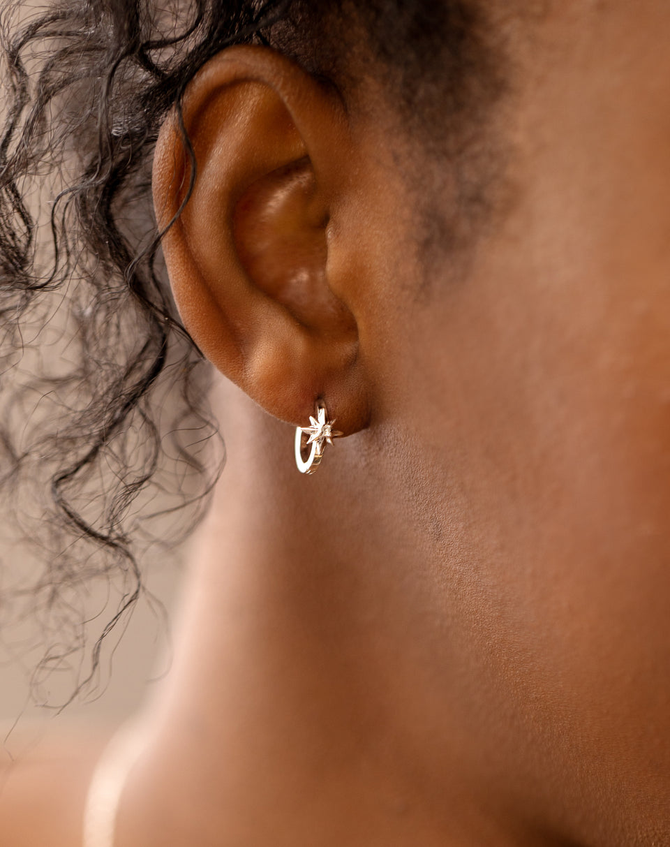 A close-up of a model wearing the North Star Hoops in silver, which features a small star charm with a central crystal.