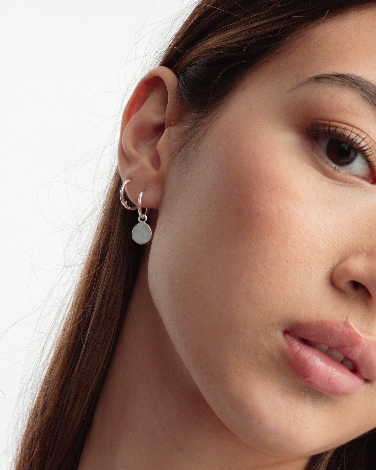 A close-up on a model wearing two Beloved Small Hoops in silver, one worn plain in the upper ear and one in the lobe styled with a round silver disc pendant.