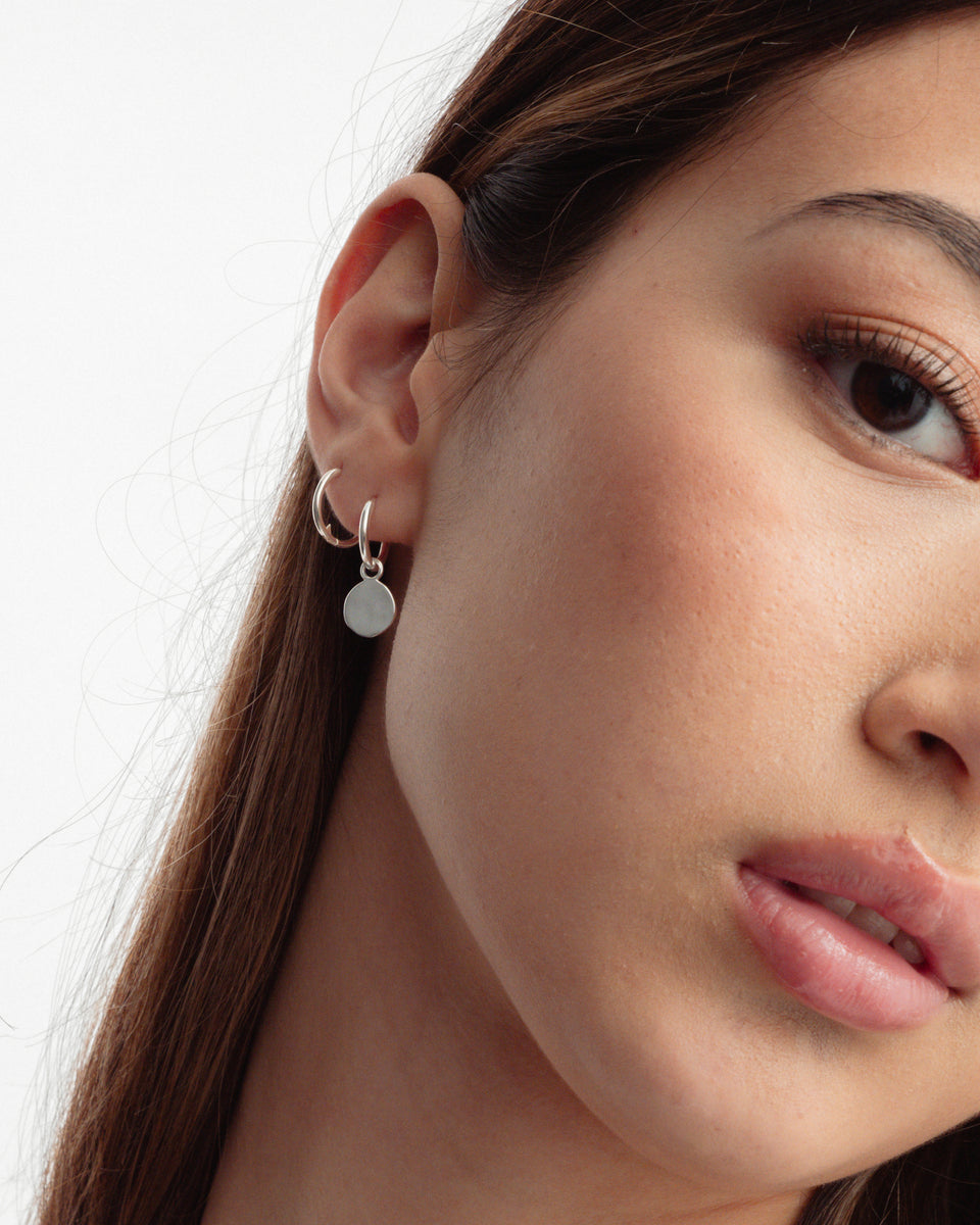 A close-up on a model wearing two Beloved Small Hoops in silver, one worn plain in the upper ear and one in the lobe styled with a round silver disc pendant.