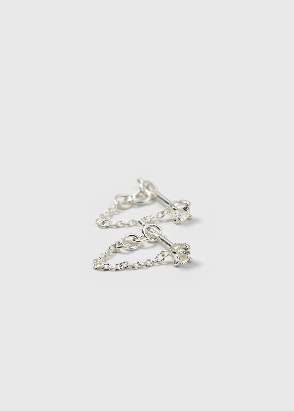 A product shot of the Minimalistica Solo Nova Earrings in silver, showing the pair of stud earrings with white topaz stones and their connecting fine silver chains.