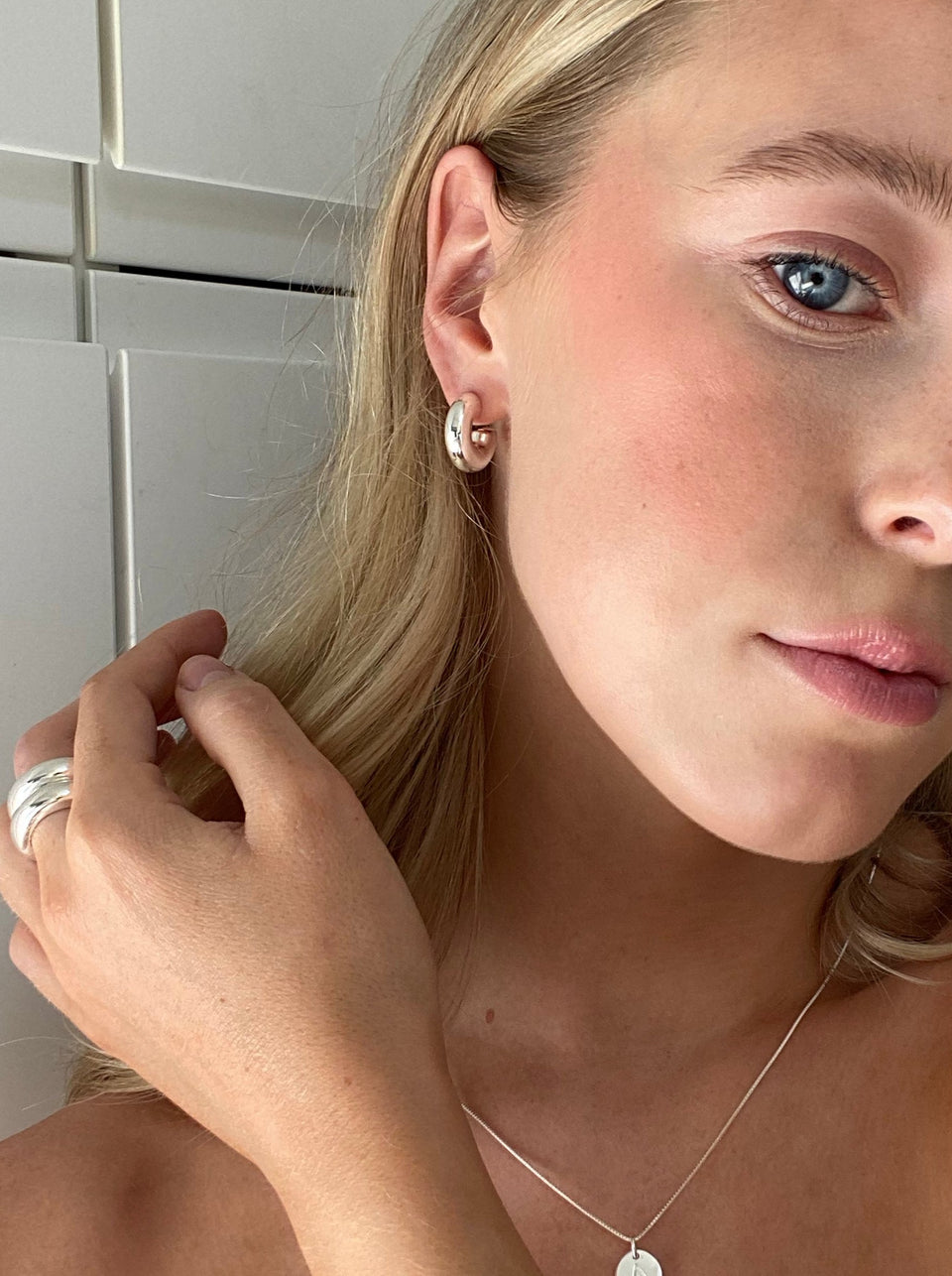 A close-up of a model wearing the chunky Bolded Little Sis Hoops in silver, paired with a matching bold silver ring and a delicate silver necklace.