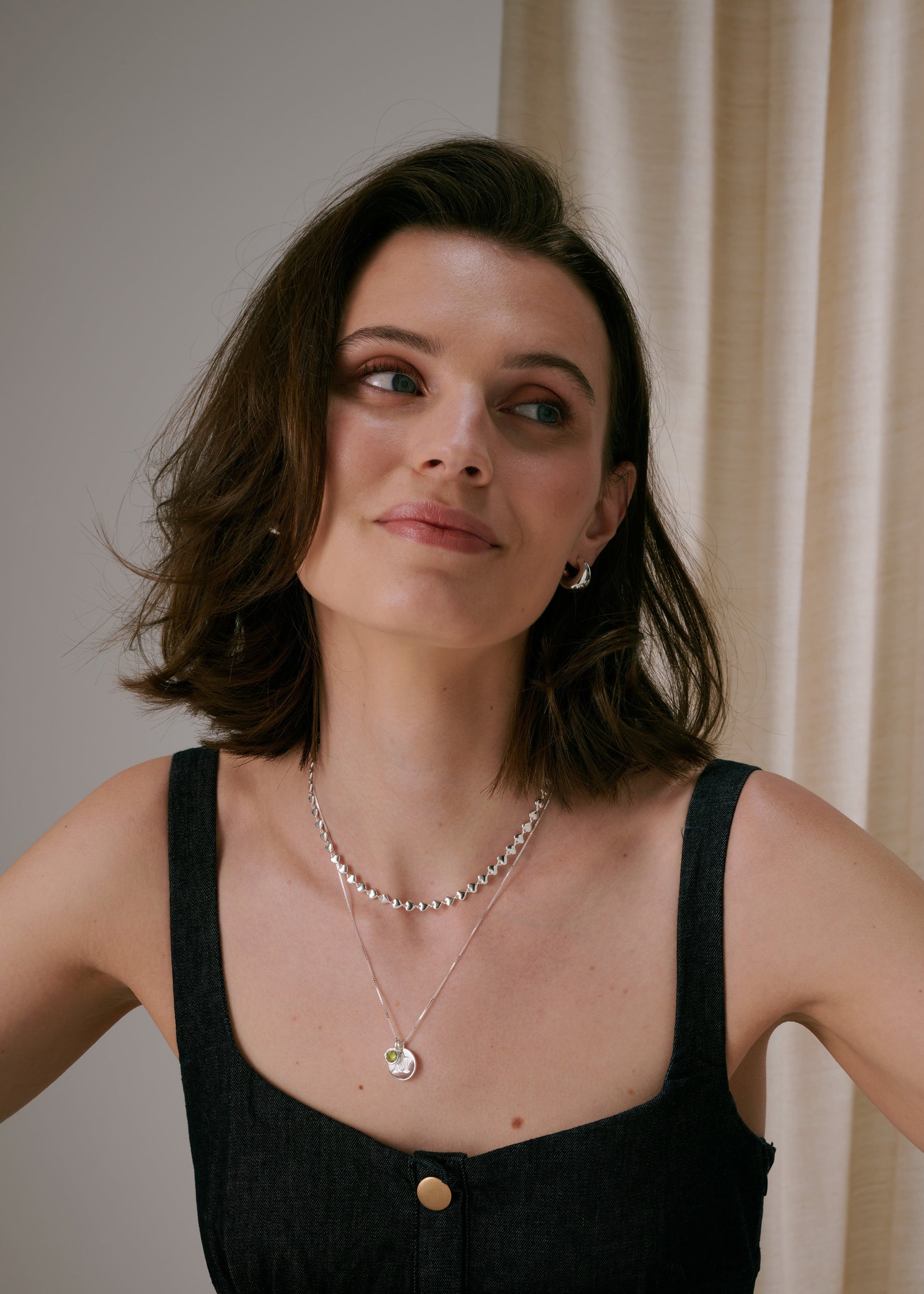 Chunky silver earrings, layered with two delicate silver necklaces: a short beaded chain and a fine chain featuring a letter pendant and the August birthstone.