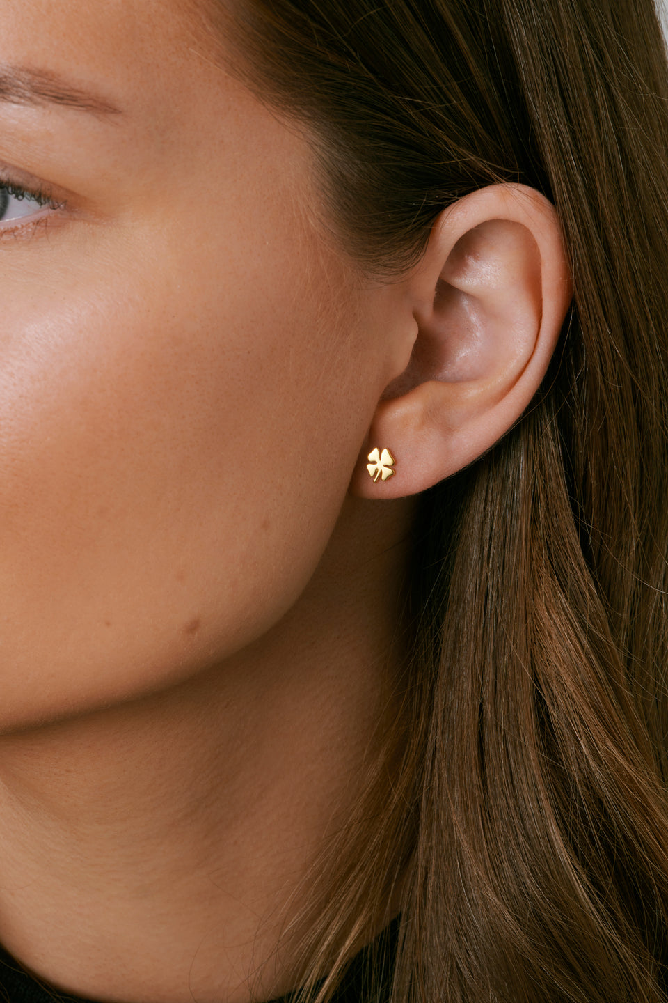 A close-up shot of a model wearing the Bring Me Luck stud earring in gold, which is shaped like a small four-leaf clover.