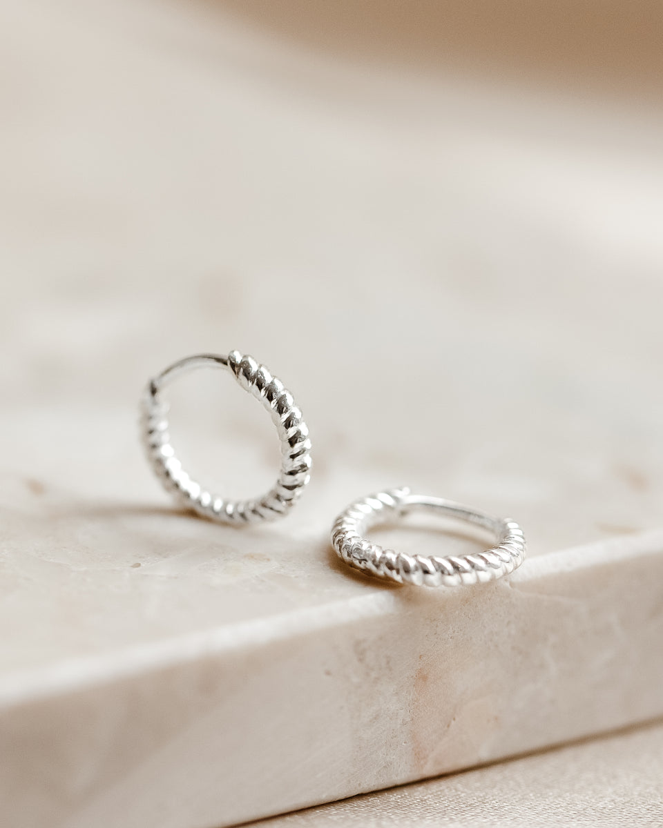 A product shot of the Beloved Twisted Small Hoops in silver, resting on a beige surface which highlights their unique twisted design.
