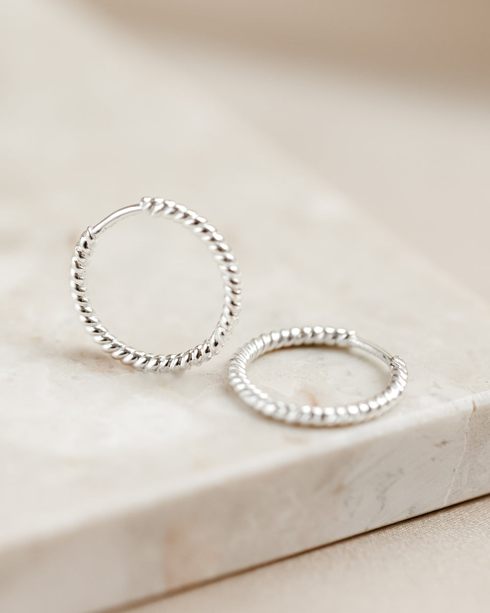 A product shot of the Beloved Twisted Medium Hoops in silver, resting on a marble surface, highlighting their intricate twisted design.