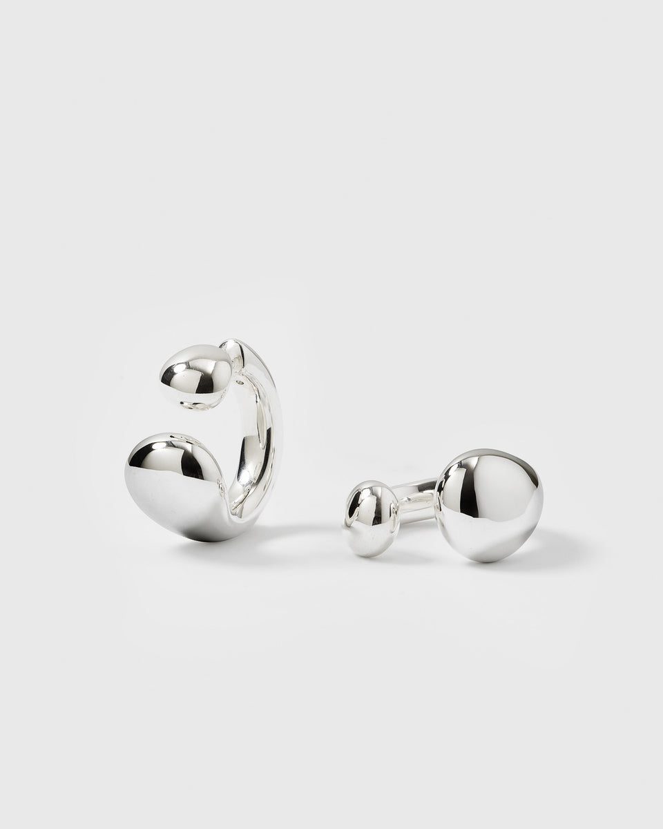 A product shot of the Bolded Drops Earrings in silver, displayed as two separate pieces to showcase their unique design: a C-shaped hoop with drop-like ends and a double-drop stud.