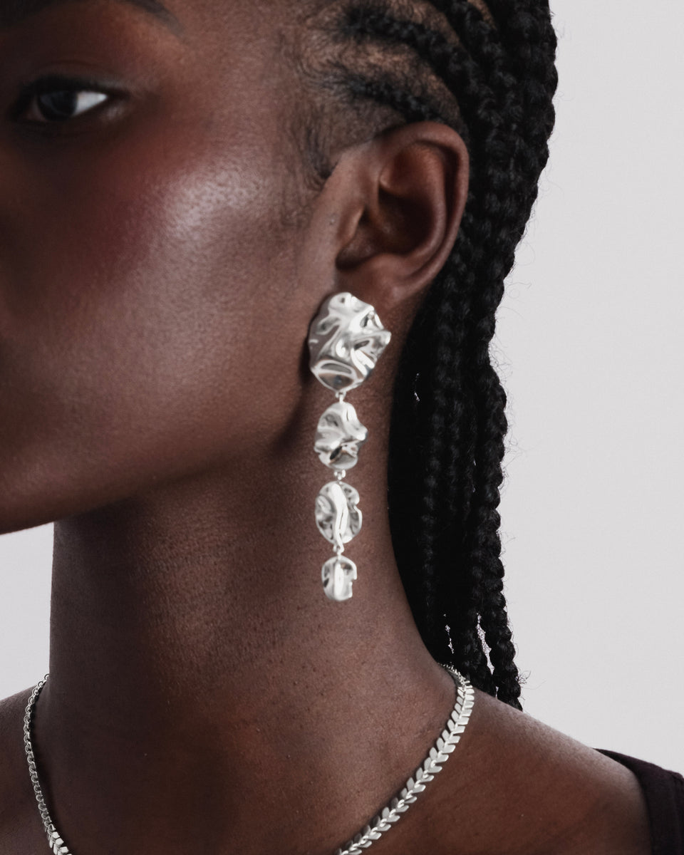 A close-up shot of a model wearing the Moulded Long Single Earring in silver. The statement earring has a long dangle design with an organic, irregular texture. She is also wearing a silver necklace with a leaf-like chain design.