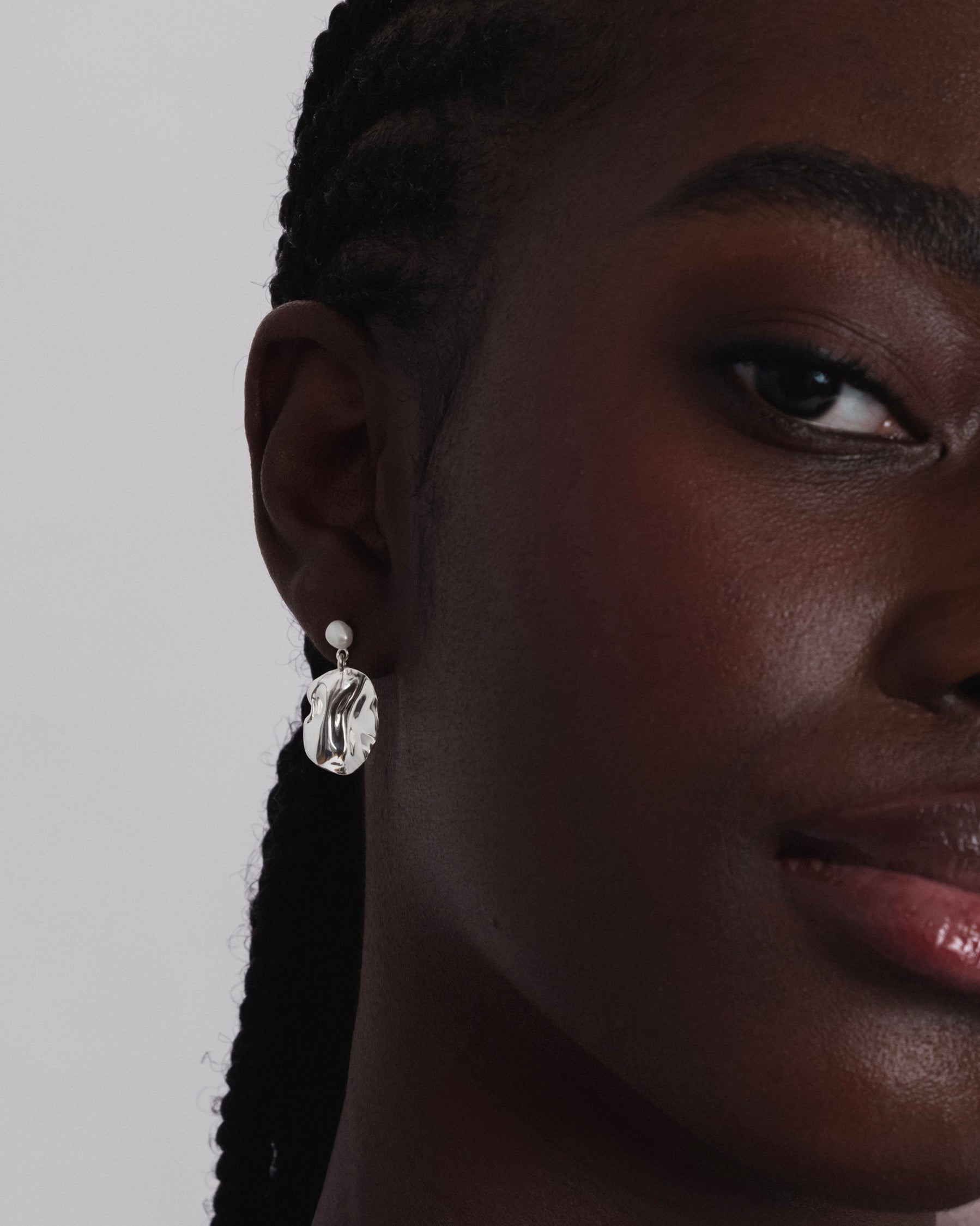 A close-up shot of a model wearing the Moulded Short Earring in silver, which features a small freshwater pearl stud and a dangling, organically shaped pendant with an irregular surface.