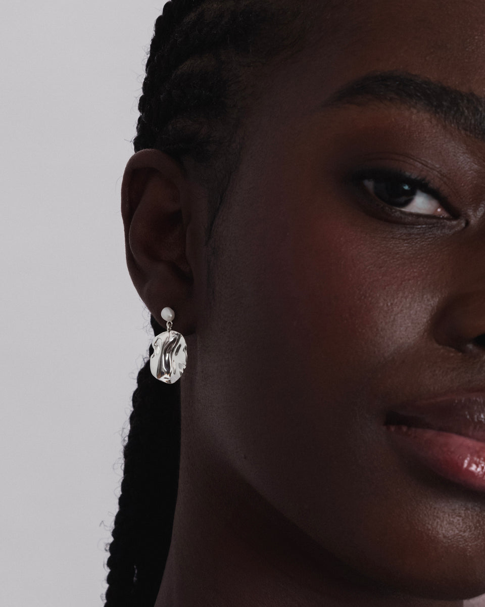 A close-up shot of a model wearing the Moulded Short Earring in silver, which features a small freshwater pearl stud and a dangling, organically shaped pendant with an irregular surface.