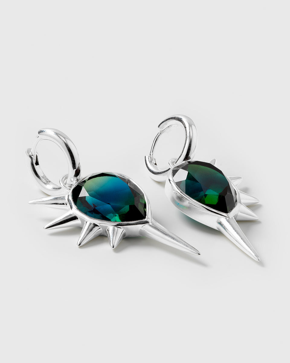 A product shot of the Queen Of Spikes Earrings in silver, featuring hoops with dangling pendants that have a large, faceted teal quartz stone and are adorned with silver spikes.