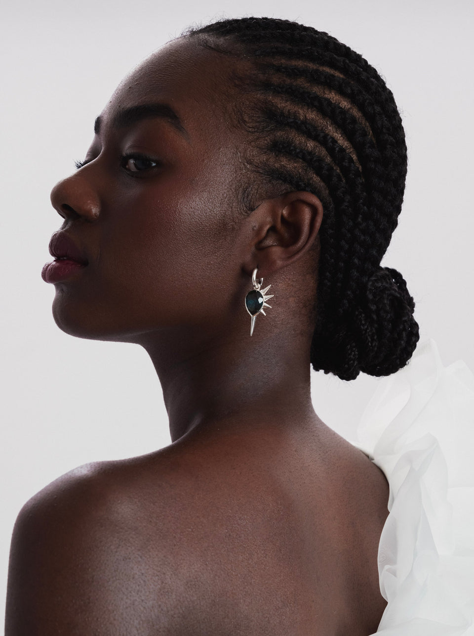 A model seen in profile wearing the Queen Of Spikes earring in silver, which features a hoop and a dangling charm with a dark teal quartz stone surrounded by spikes.