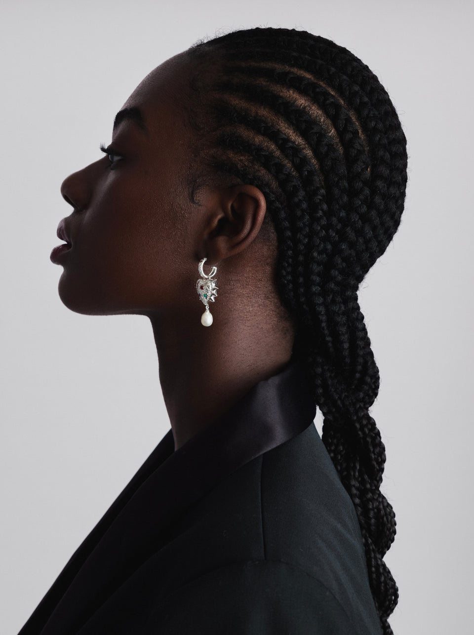 A close-up, side profile shot of a model wearing the Drama Queen Heart Pearl Earrings in silver, featuring a heart-shaped charm with spikes, three colored stones, and a dangling freshwater pearl.
