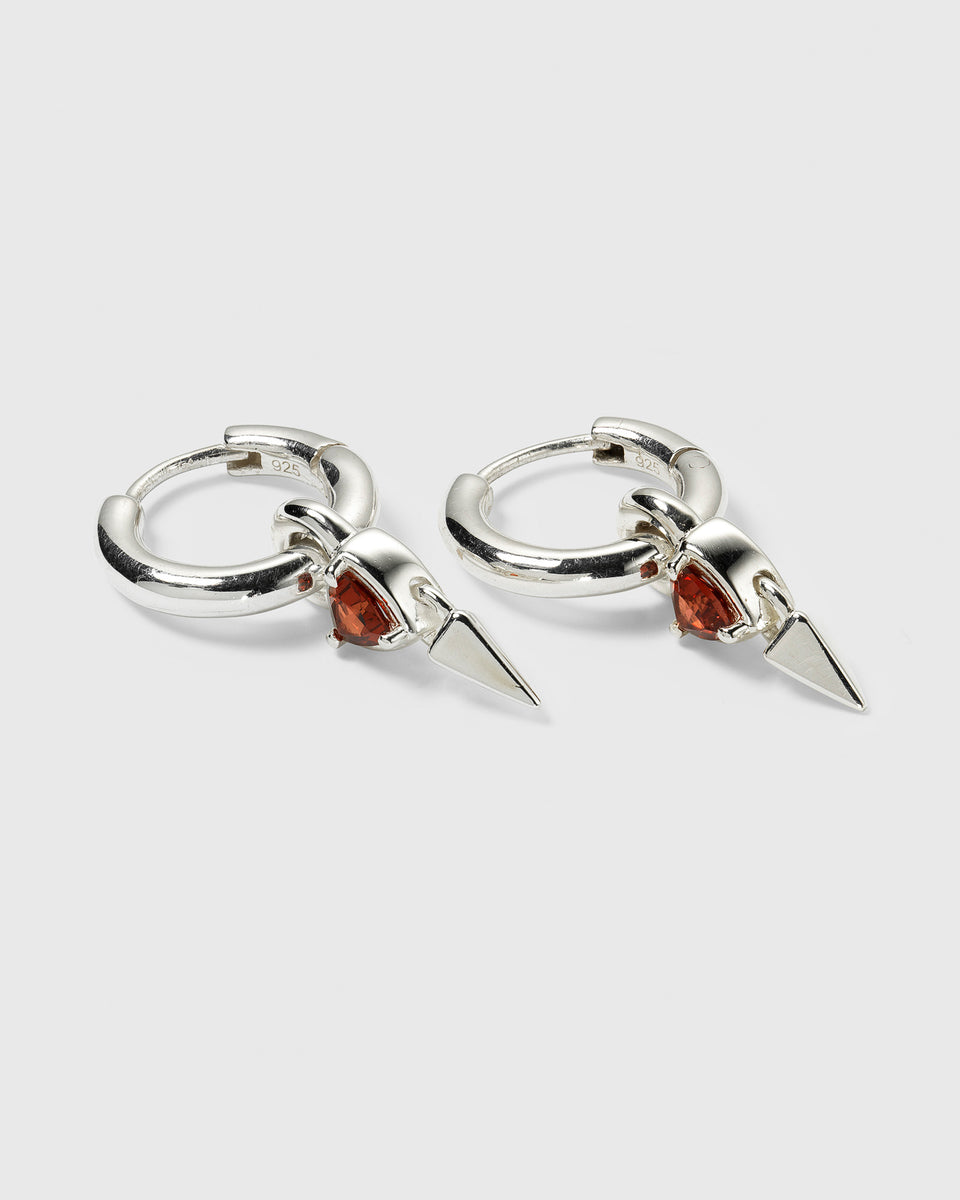 A product shot of the Mrs H Spike Earrings in silver, featuring chunky hoops with dangling charms, each adorned with a red garnet and a pointed spike.
