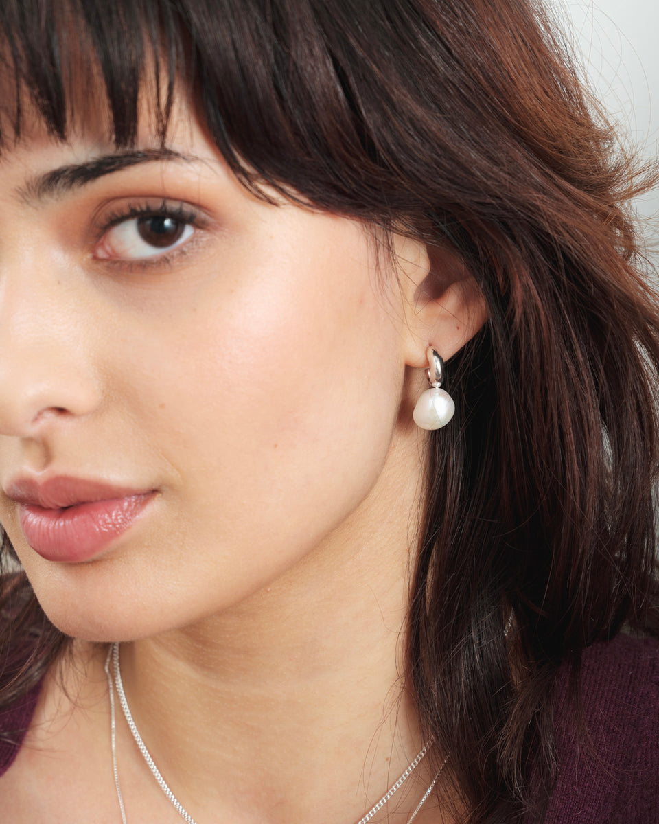 A close-up shot of a model wearing the silver Paxton earring with a baroque pearl. Two delicate silver necklaces is also visible.