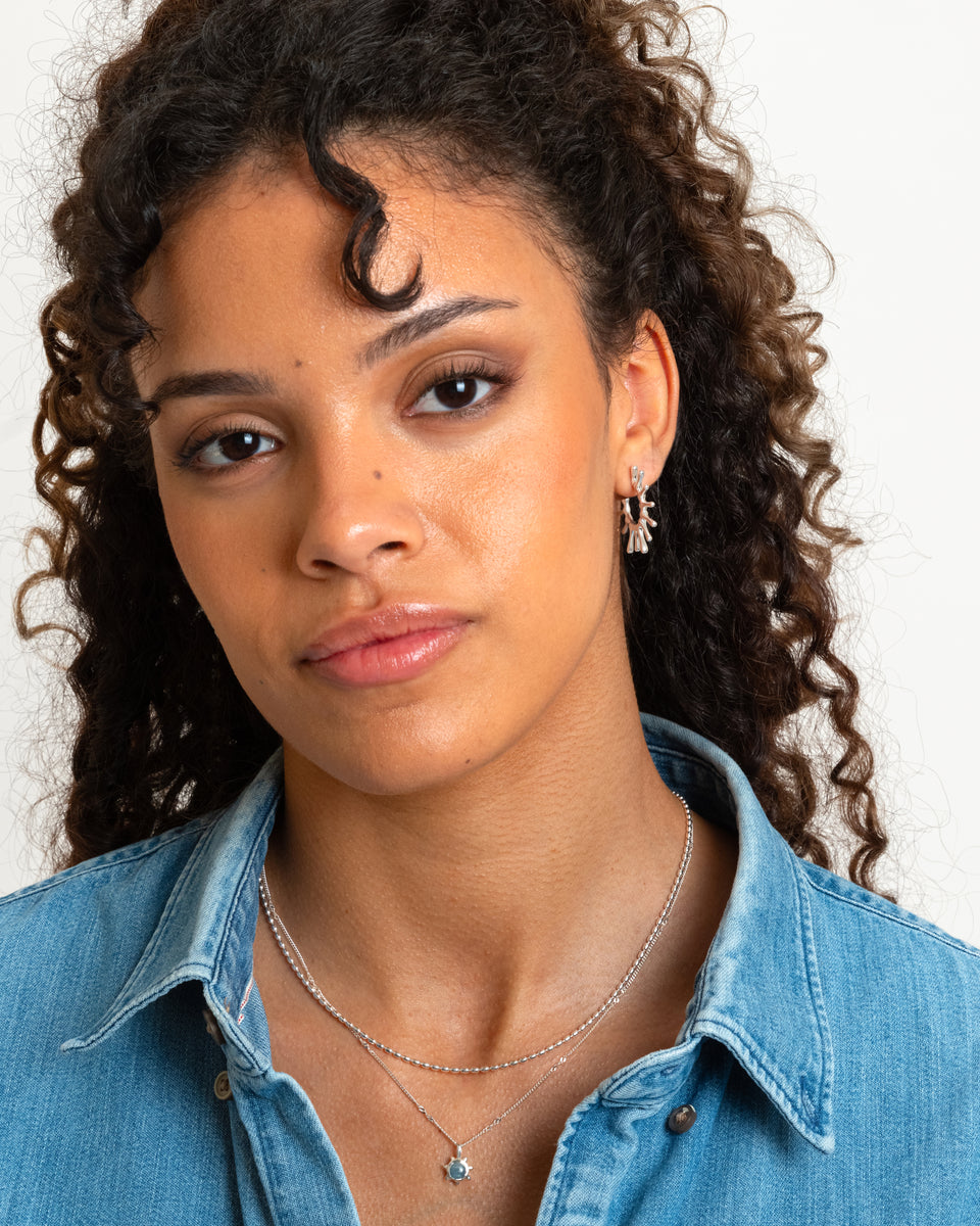 A close-up shot of a model wearing the Florentina Big Earrings in silver, which feature a unique sun-inspired design, paired with two layered silver necklaces.
