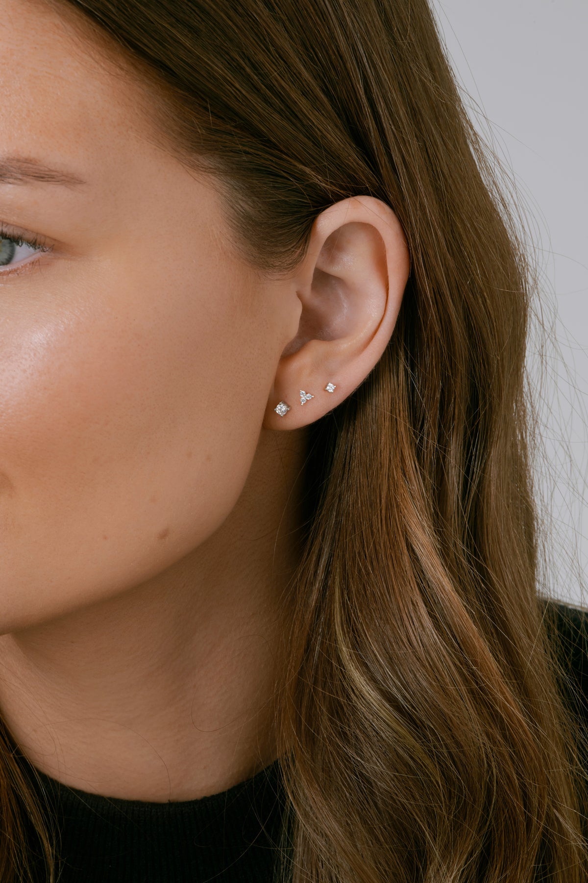 A close-up shot of a model wearing the Celestia Triple Stone Studs in silver, featuring a cluster of three clear stones. They are styled alongside two smaller, single-stone silver studs in her earlobe.