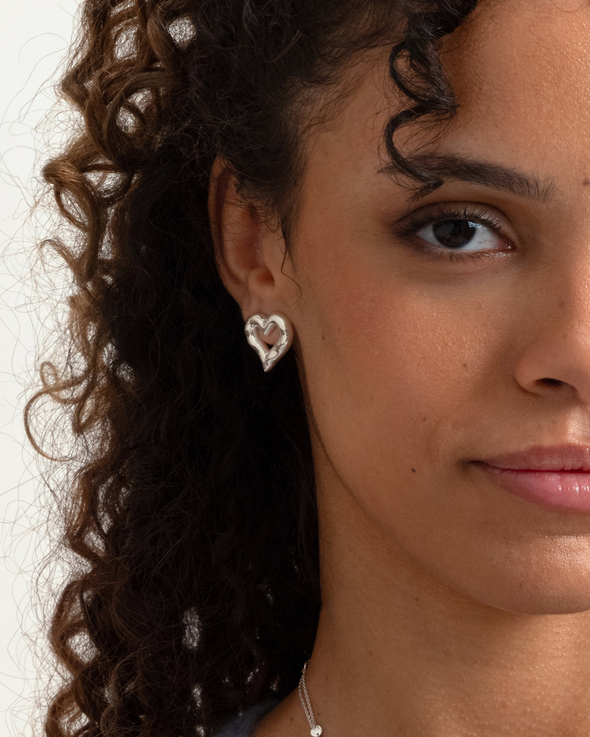 A close-up of a model wearing the Puff Heart Earring in silver, which has a chunky, organic shape. A fine silver chain necklace is also visible.