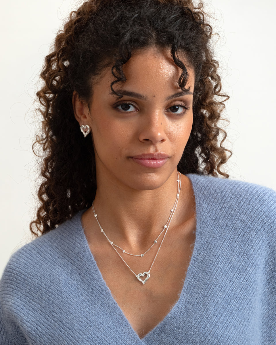 A model wears the silver Puff Heart Earrings, styled with a matching layered necklace set that includes a silver Puff Heart pendant and a delicate chain adorned with small, round hammered circles.