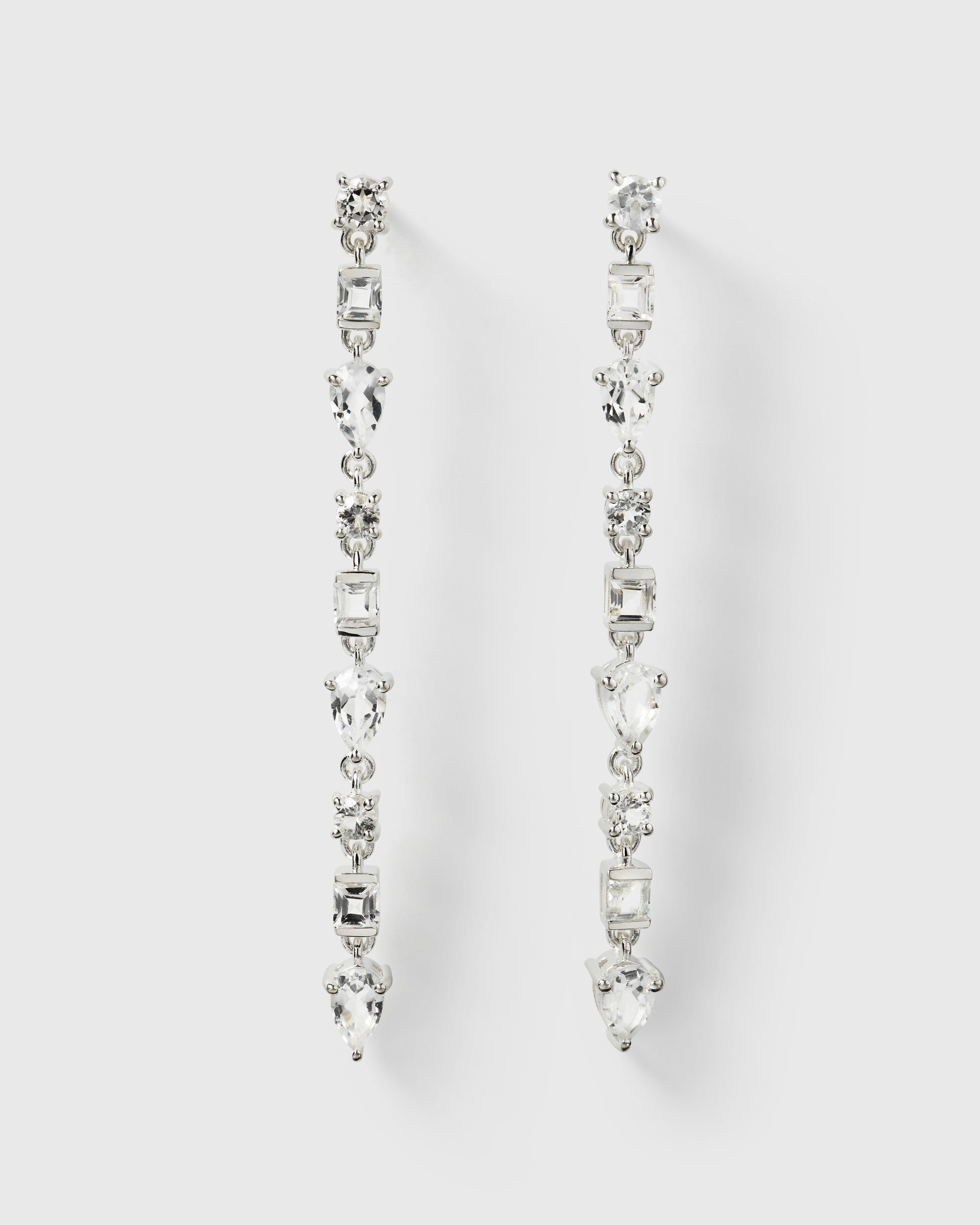 A product shot of the Pam Long Earrings in silver, laid out to display their long design featuring a pattern of sparkling white topaz stones in different shapes.