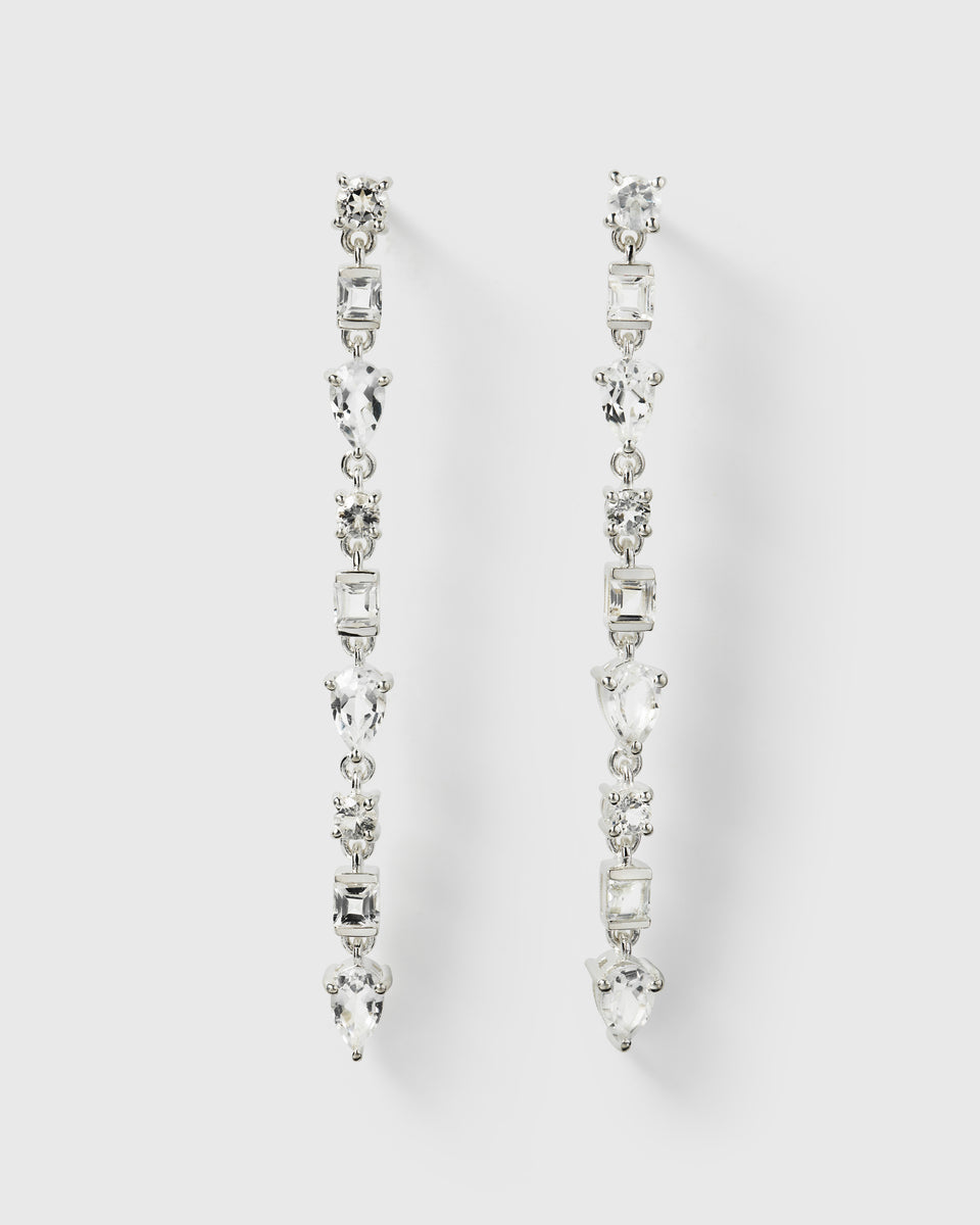 A product shot of the Pam Long Earrings in silver, laid out to display their long design featuring a pattern of sparkling white topaz stones in different shapes.