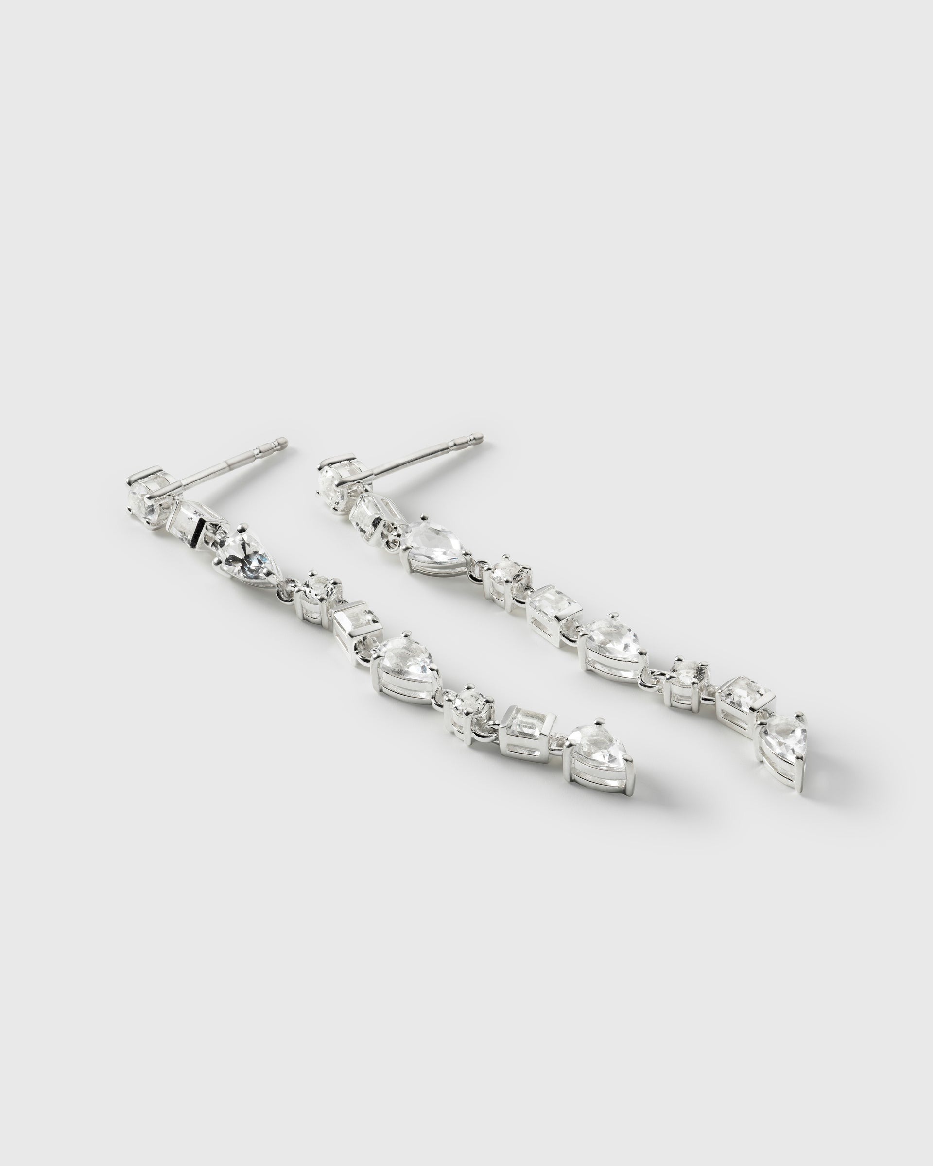 A product shot of the Pam Long Earrings in silver, laid out to showcase their elegant dangling design of interconnected white topazes in various shapes.