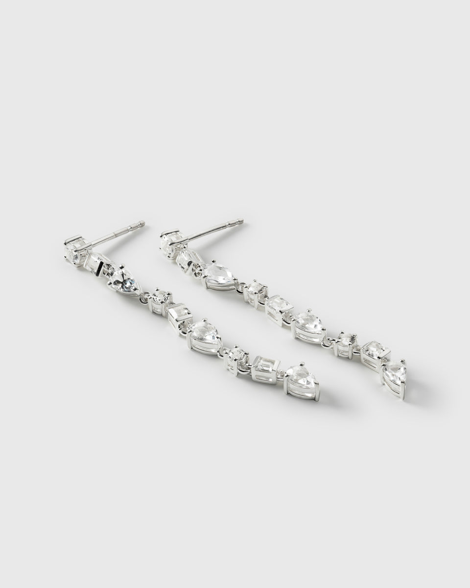 A product shot of the Pam Long Earrings in silver, laid out to showcase their elegant dangling design of interconnected white topazes in various shapes.