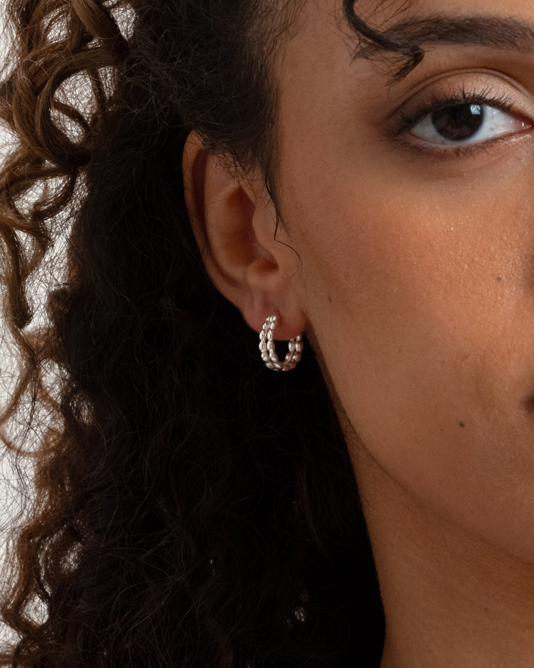 A close-up shot of a model wearing the Lunetta Earrings in silver, showcasing their unique design of two parallel hoops joined by a pattern of small ovals.