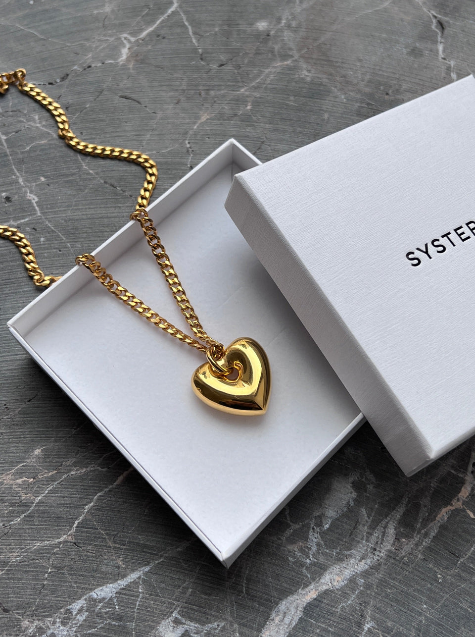 The Fat Heart Necklace in gold, featuring a chunky chain and a large, puffy heart-shaped pendant, resting inside a white Syster P jewelry box.