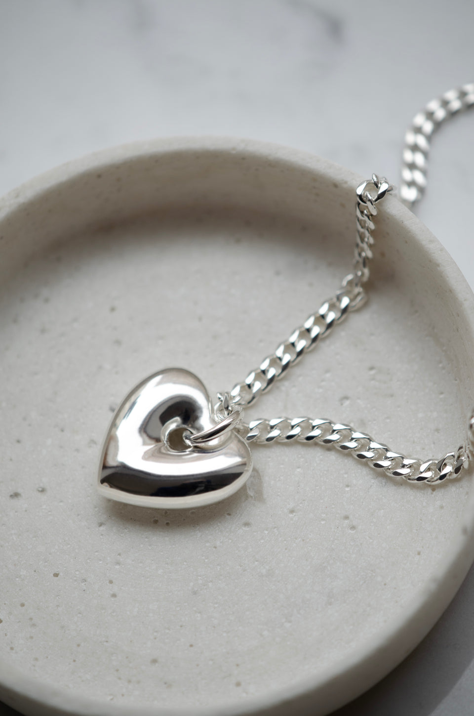 A close-up shot of the Fat Heart Necklace in silver, displayed in a light-colored dish, highlighting its chunky chain and large, puffy heart pendant.