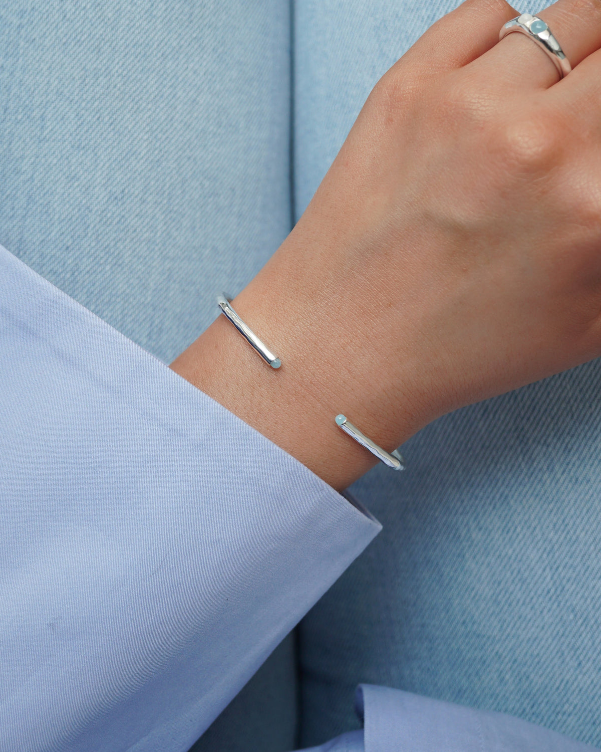 A model's wrist wearing the Florentina Bangle in silver, featuring a hammered texture and light blue aquamarine stones at the ends. She is also wearing a matching silver ring.