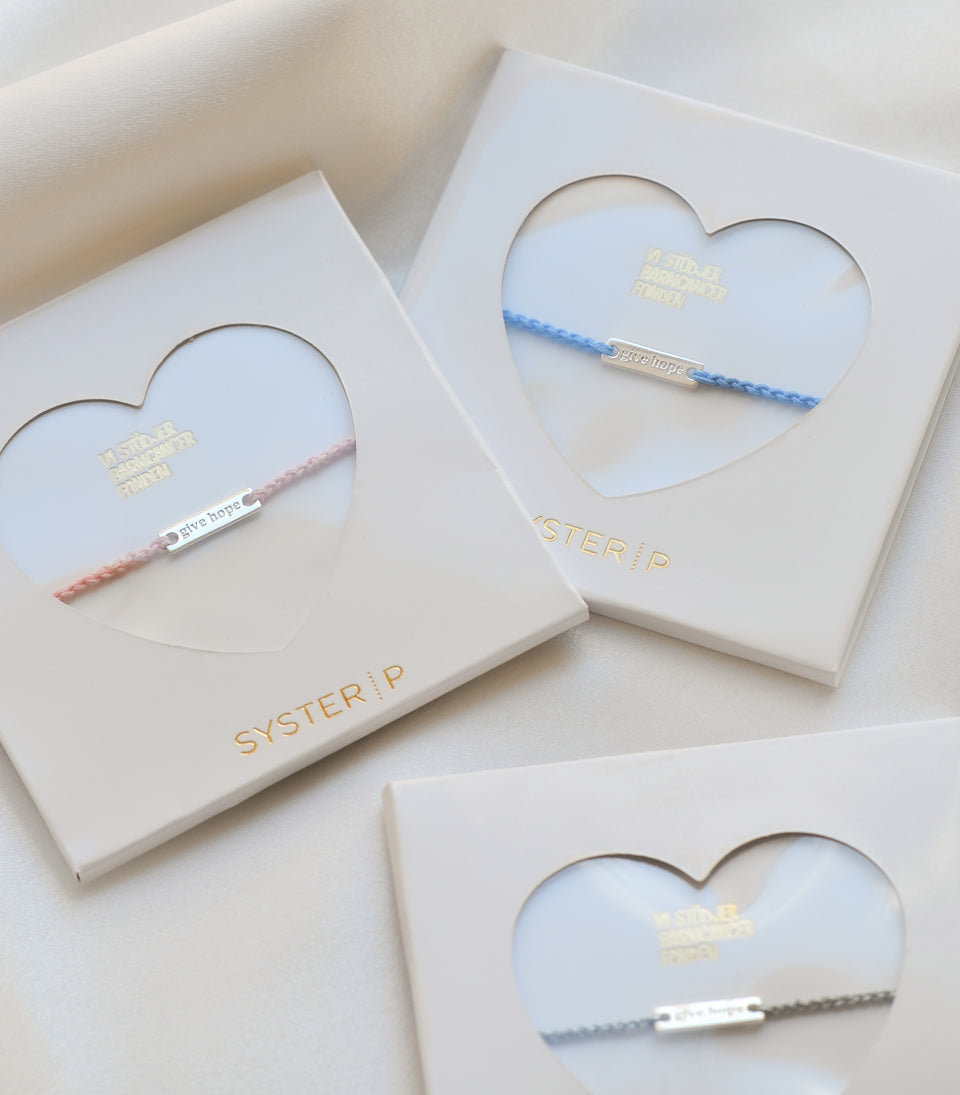 A product shot of three Give Hope thread bracelets in pink, blue, and grey, each displayed in a white Syster P box with a heart-shaped window, showcasing the silver plaque engraved with 'give hope'.
