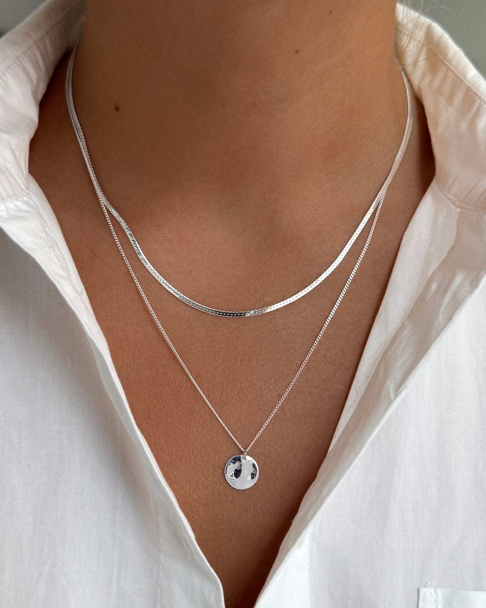 A close-up of a model wearing the Herringbone Necklace Silver, layered with the Minimalistica Hammered Necklace Silver, which features a fine chain and a round, hammered pendant.