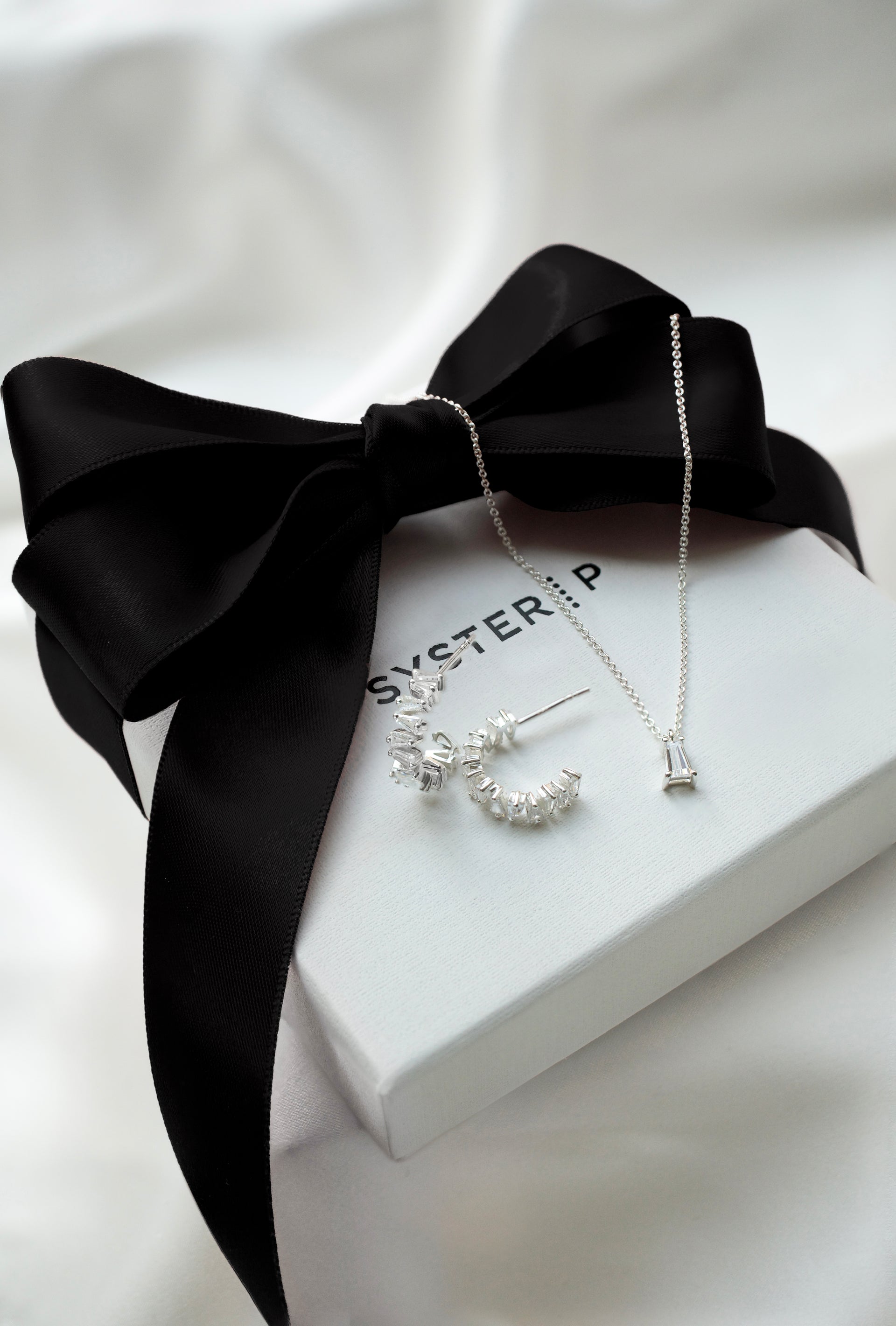 A product shot of the Iggy Earrings and matching Iggy Necklace in silver, both featuring baguette-cut cubic zirconias, displayed on a white Syster P gift box tied with a black ribbon.