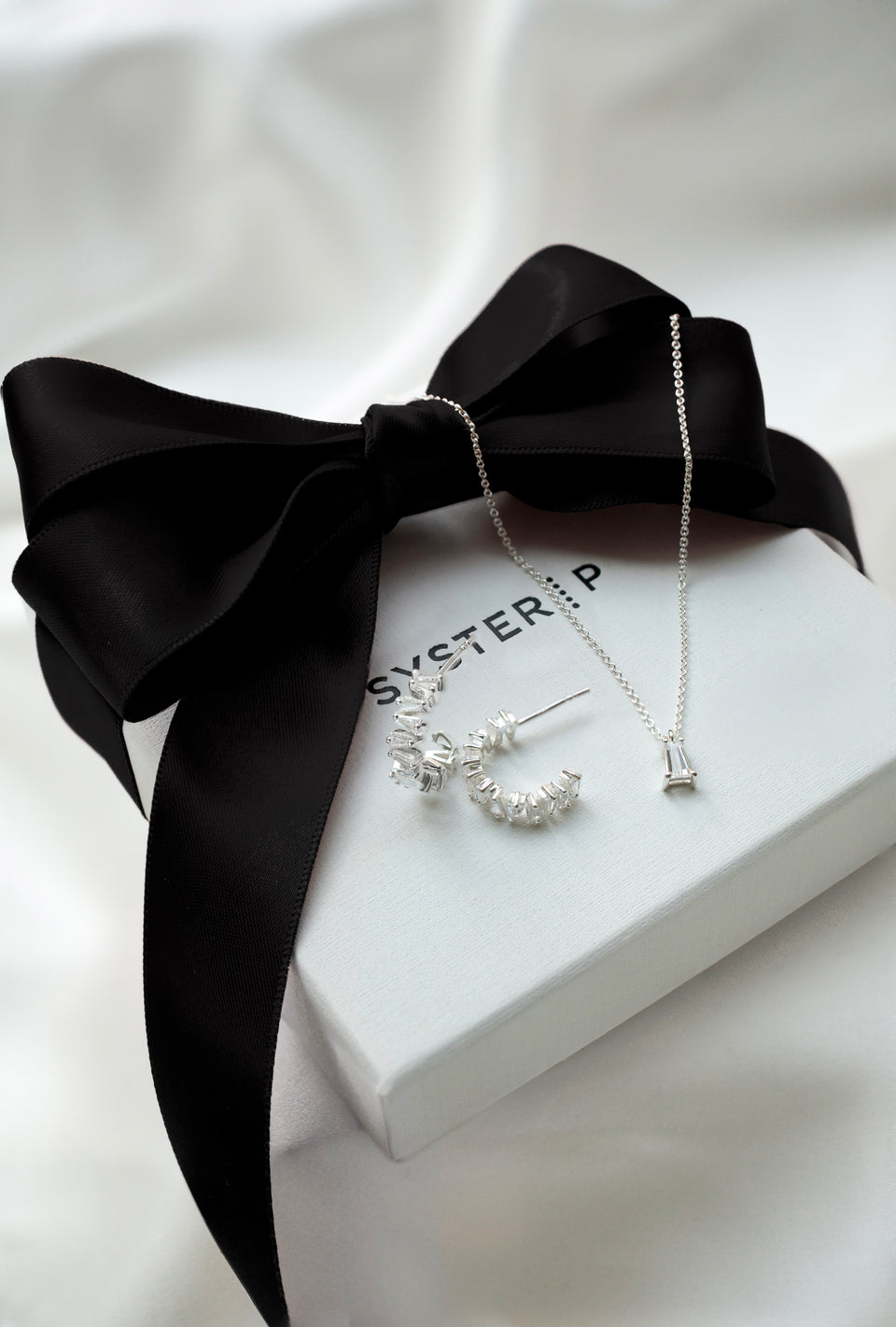 A product shot of the Iggy Earrings and matching Iggy Necklace in silver, both featuring baguette-cut cubic zirconias, displayed on a white Syster P gift box tied with a black ribbon.