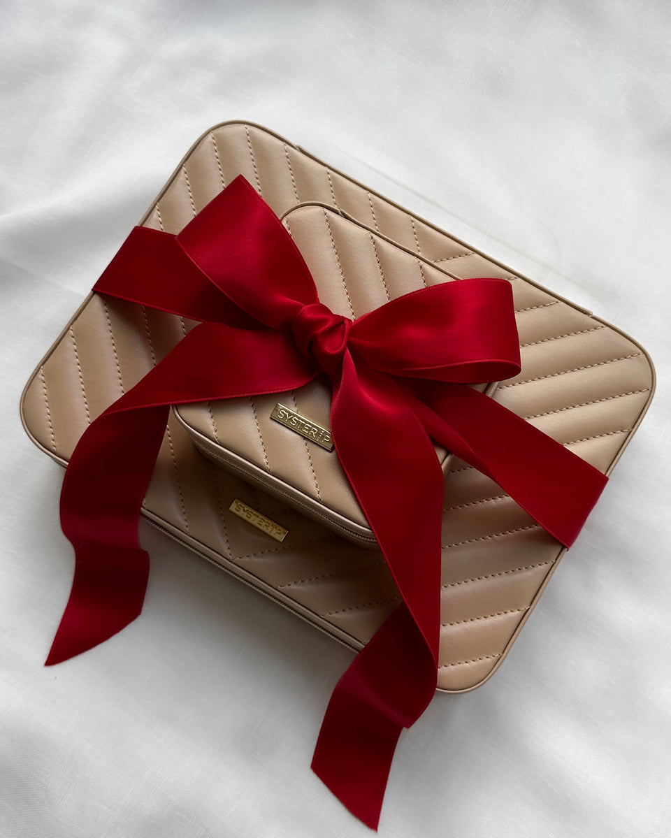 A large and a small beige Syster P jewelry case with a quilted pattern, stacked on top of each other and tied with a large, red satin bow.