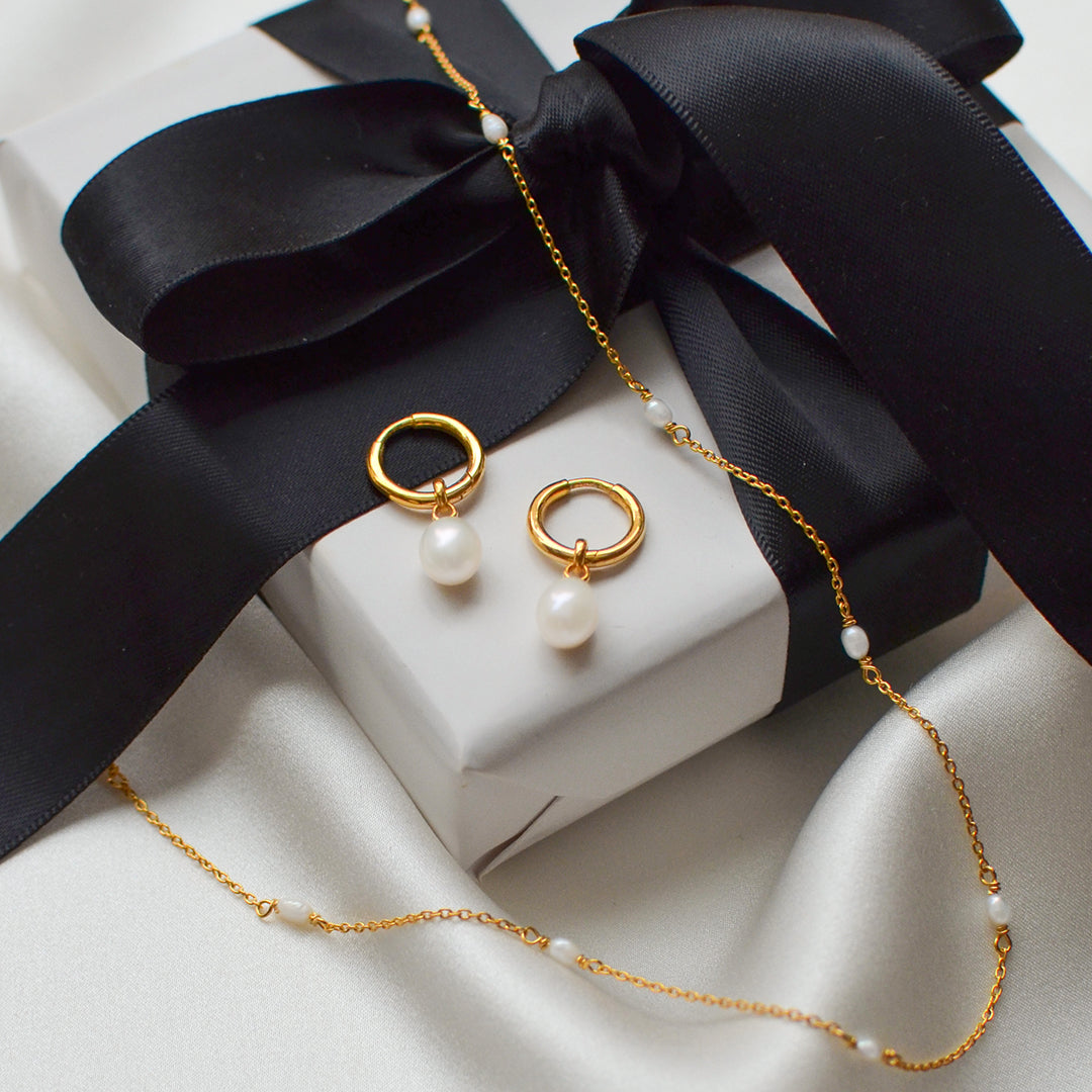 The Treasure Necklace & Earrings Set in gold, featuring gold hoop earrings with dangling freshwater pearls and a matching necklace, elegantly displayed on a white gift box with a black ribbon.