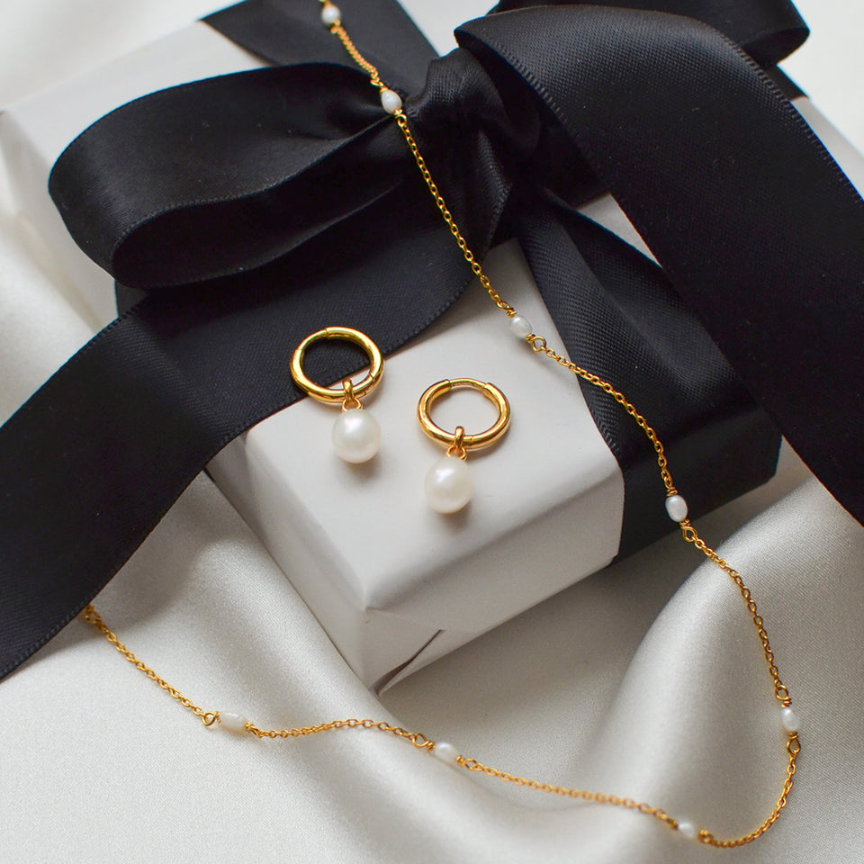 The Treasure Necklace & Earrings Set in gold, featuring gold hoop earrings with dangling freshwater pearls and a matching necklace, elegantly displayed on a white gift box with a black ribbon.