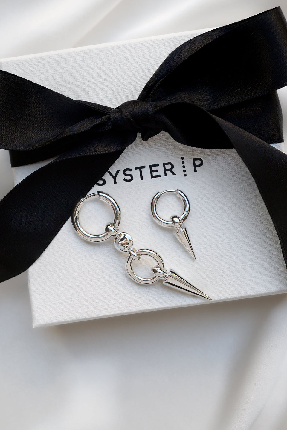 A product shot of the Juno Long Earring and the Juno Single Hoop Earring in silver, displayed on a white Syster P gift box to highlight their edgy design with dangling spikes.