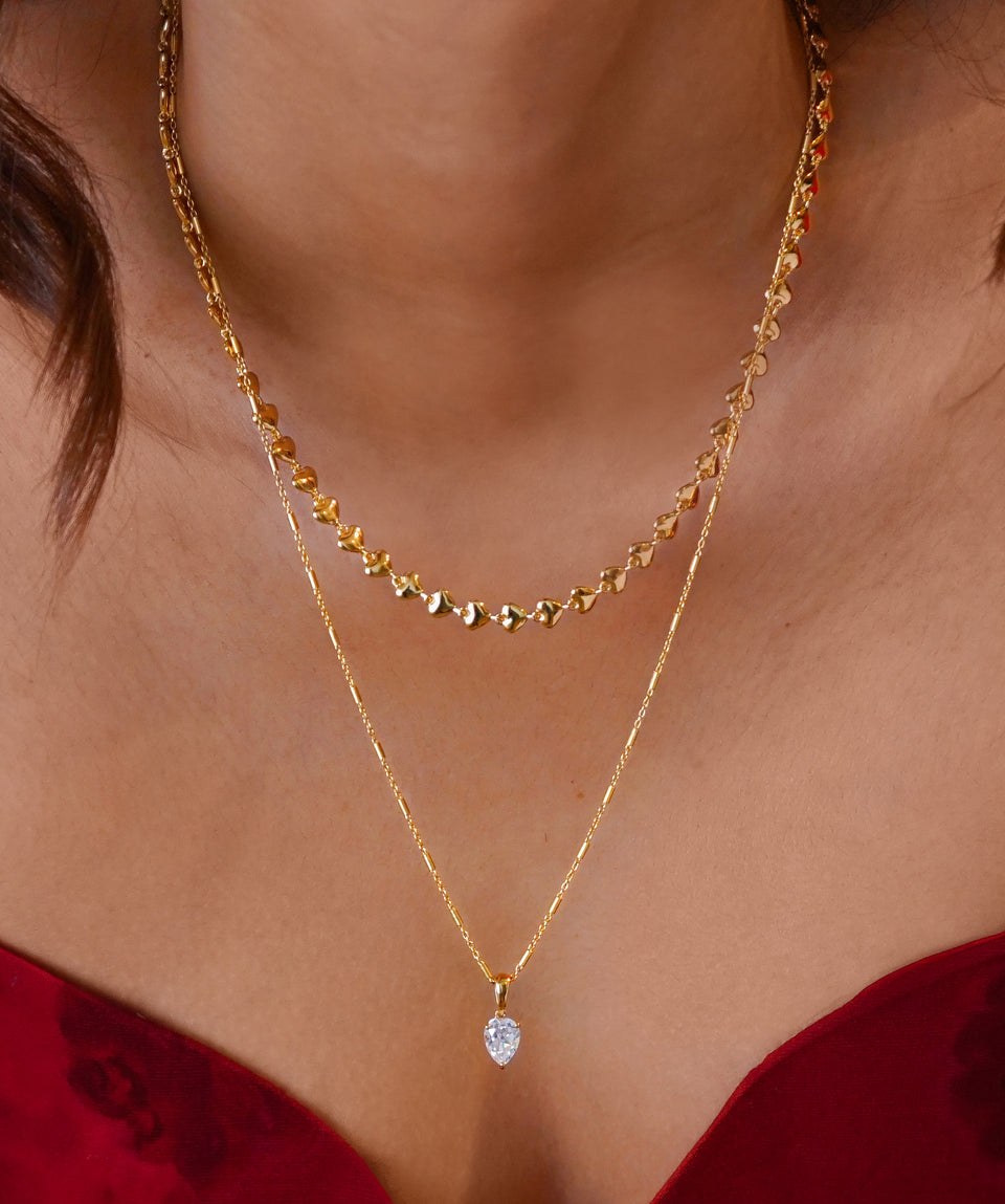 A close-up of a model wearing the Darling Necklace in gold, a chain made of small hearts, layered with the Katie Necklace, a longer gold chain featuring a pear-shaped crystal pendant.