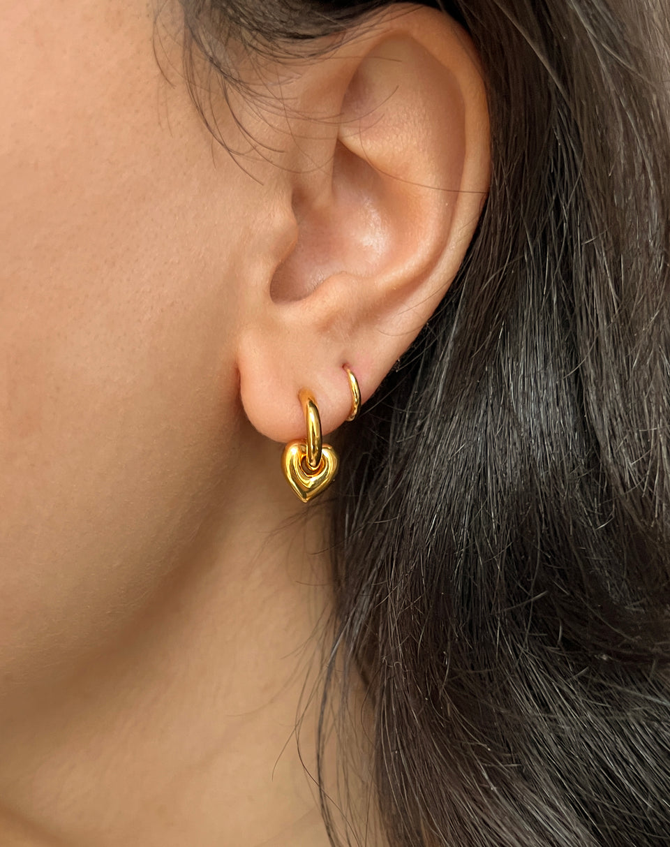 A close-up of a model wearing the Beloved Mini Hoops in gold, styled with another gold hoop earring featuring a dangling, puffy heart charm.