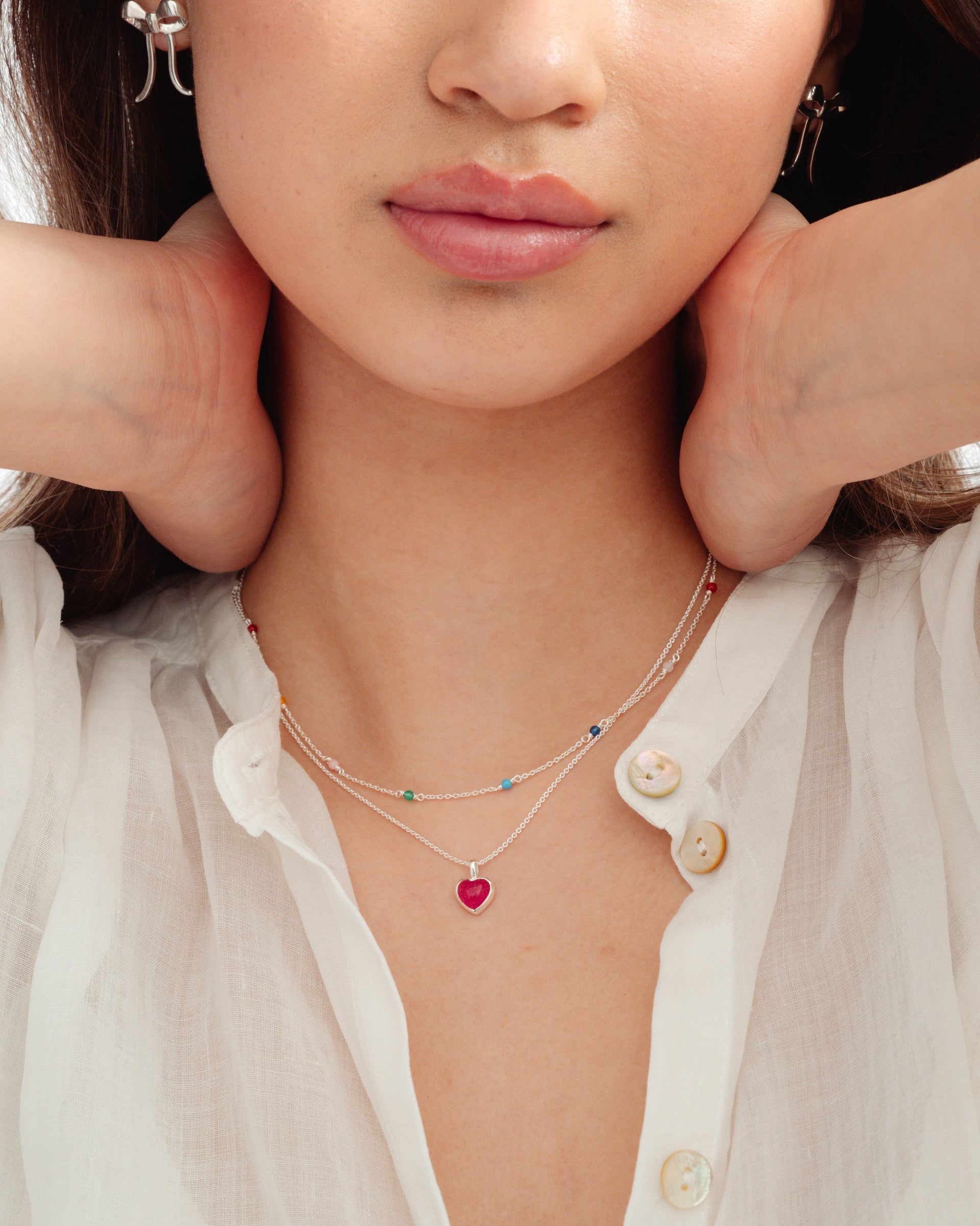 Close-up of a model wearing the silver La La Love Necklace featuring a pink heart pendant, layered with the Santa Monica Necklace with colourful beads. She is also wearing the silver bow-shaped Rosie Earrings.